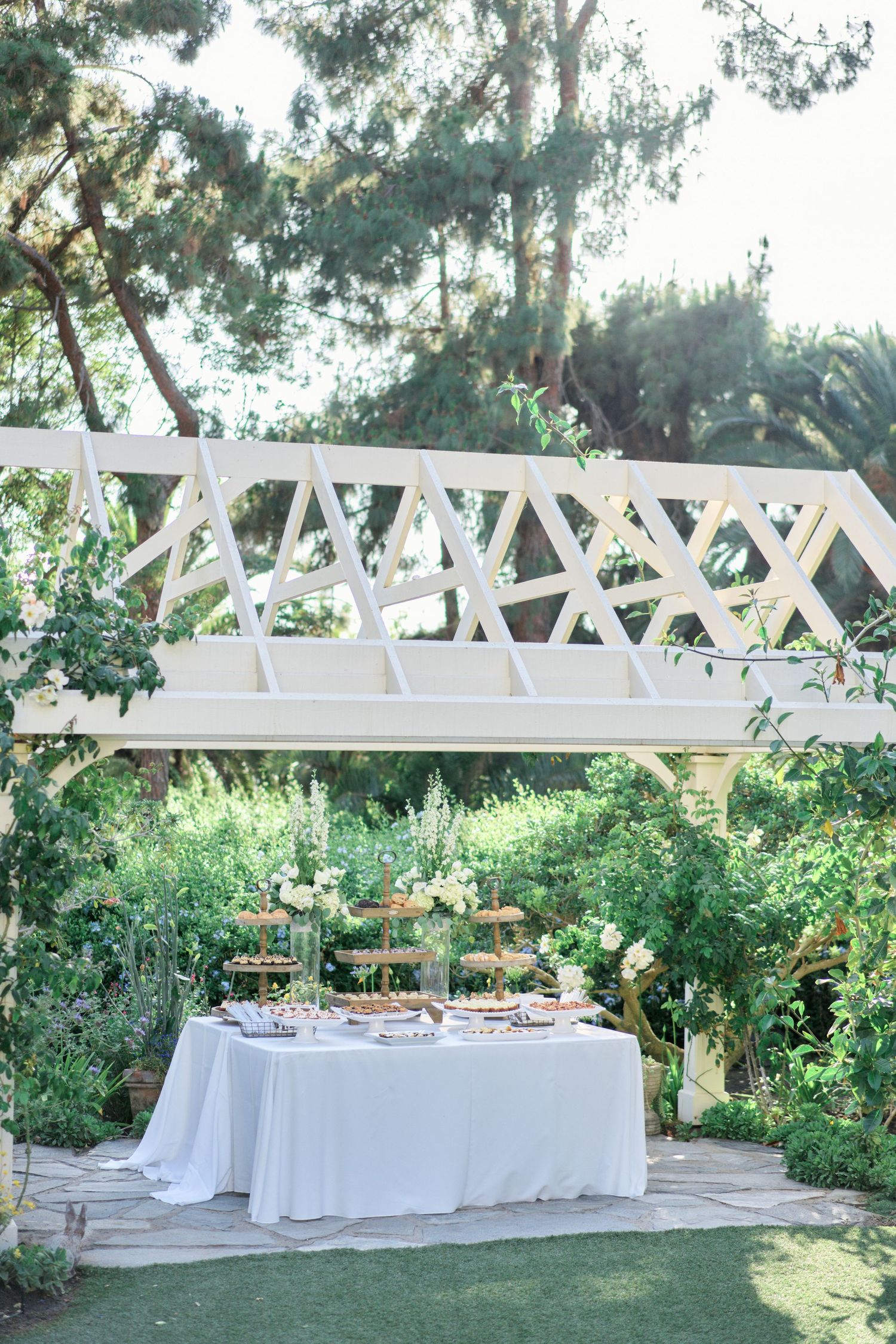 White reception table set beneath a geometric garden trellis with natural greenery surroundings.