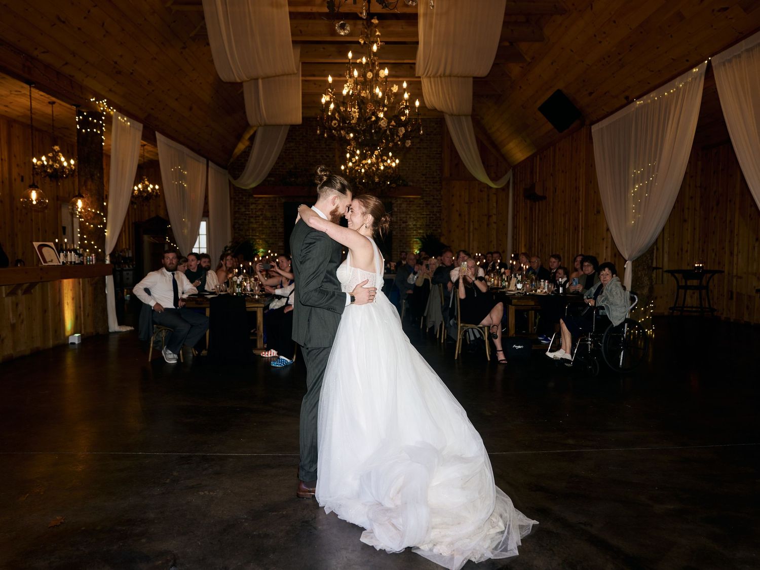 A couple shares their first dance in an elegant wooden reception hall with chandeliers and white draping.