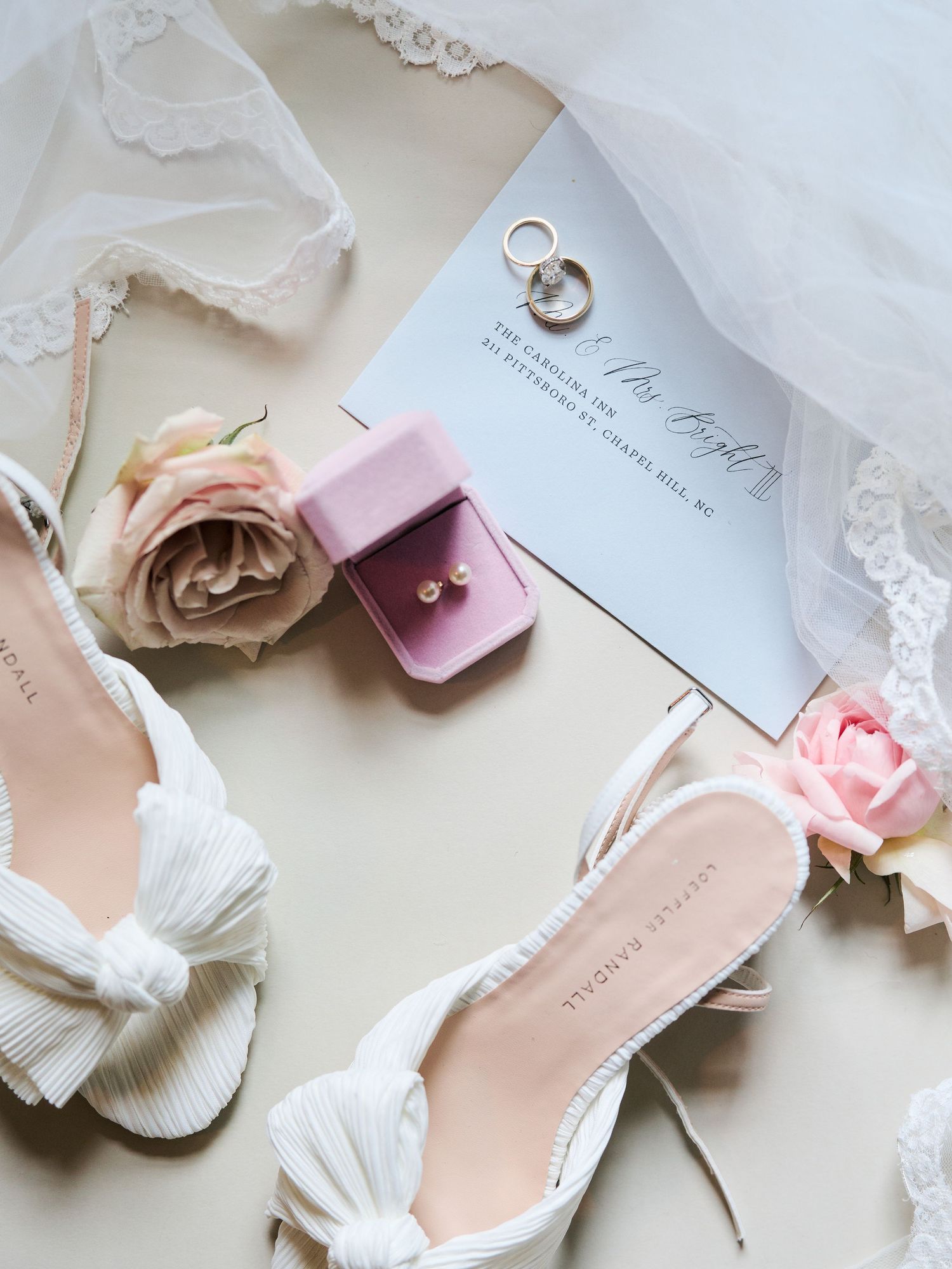 Elegant bridal accessories including white shoes, blush roses, and delicate lace details arranged on a light background.
