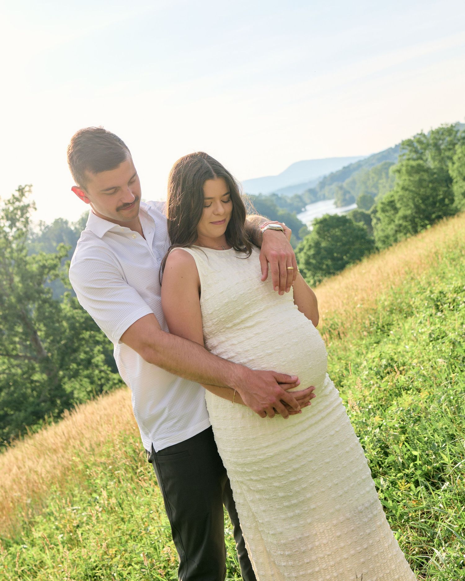 A couple shares a tender maternity moment outdoors, man embracing pregnant woman in white dress on a scenic hillside.