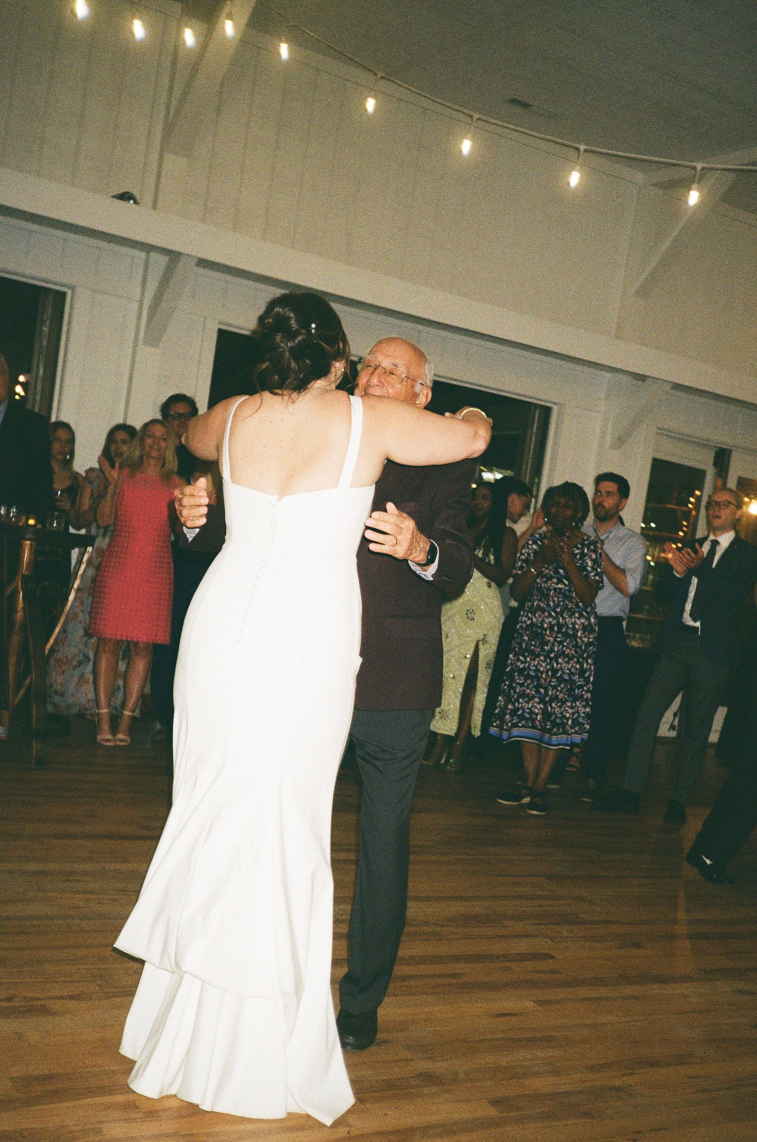 Bride in white gown shares first dance with partner at elegant wedding reception with string lights.