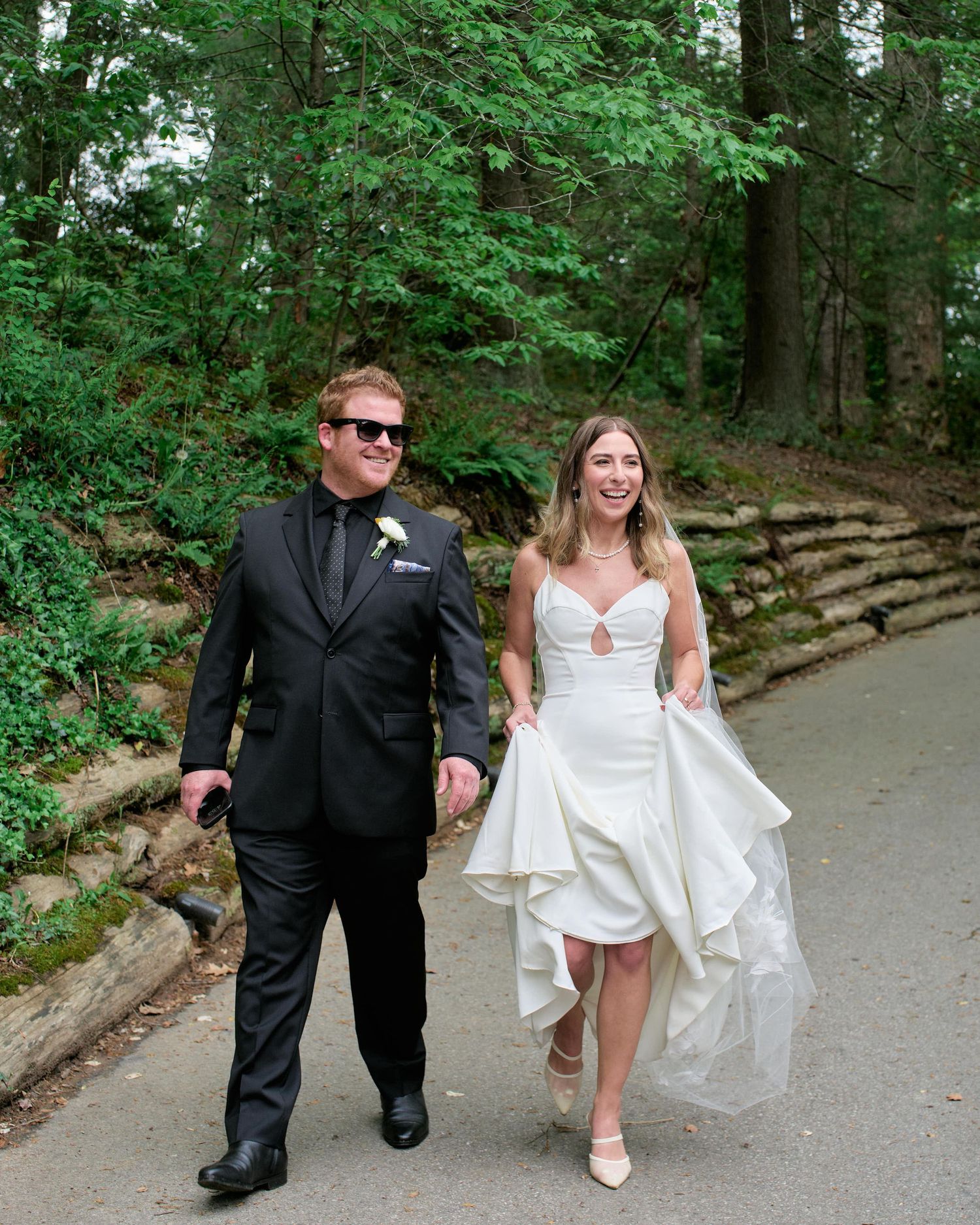 A couple walking together on a wooded path, with the person in a black suit and another in a white dress.