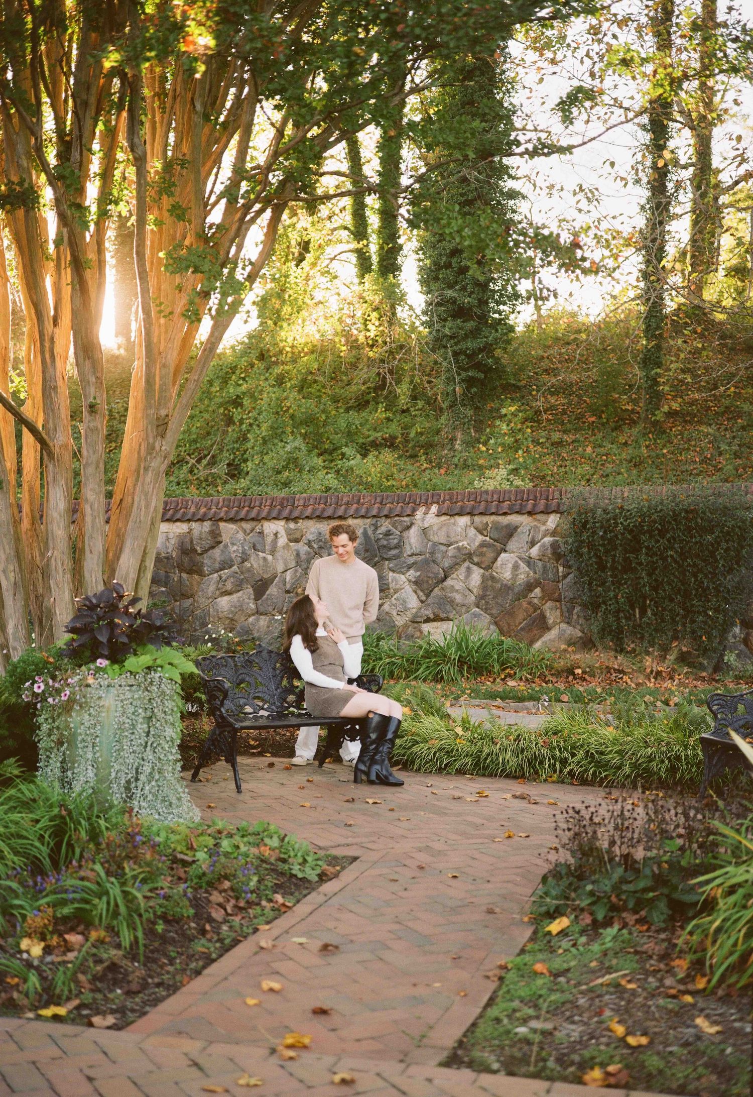 Couple sitting on iron bench in sunlit garden with brick path, stone wall, and ivy-covered trees in autumn.