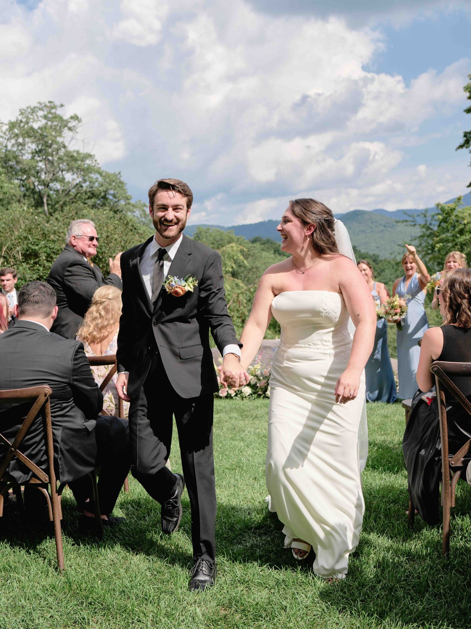 Joyful newlyweds walk down outdoor aisle holding hands, groom in black tuxedo, bride in strapless gown, mountain backdrop.