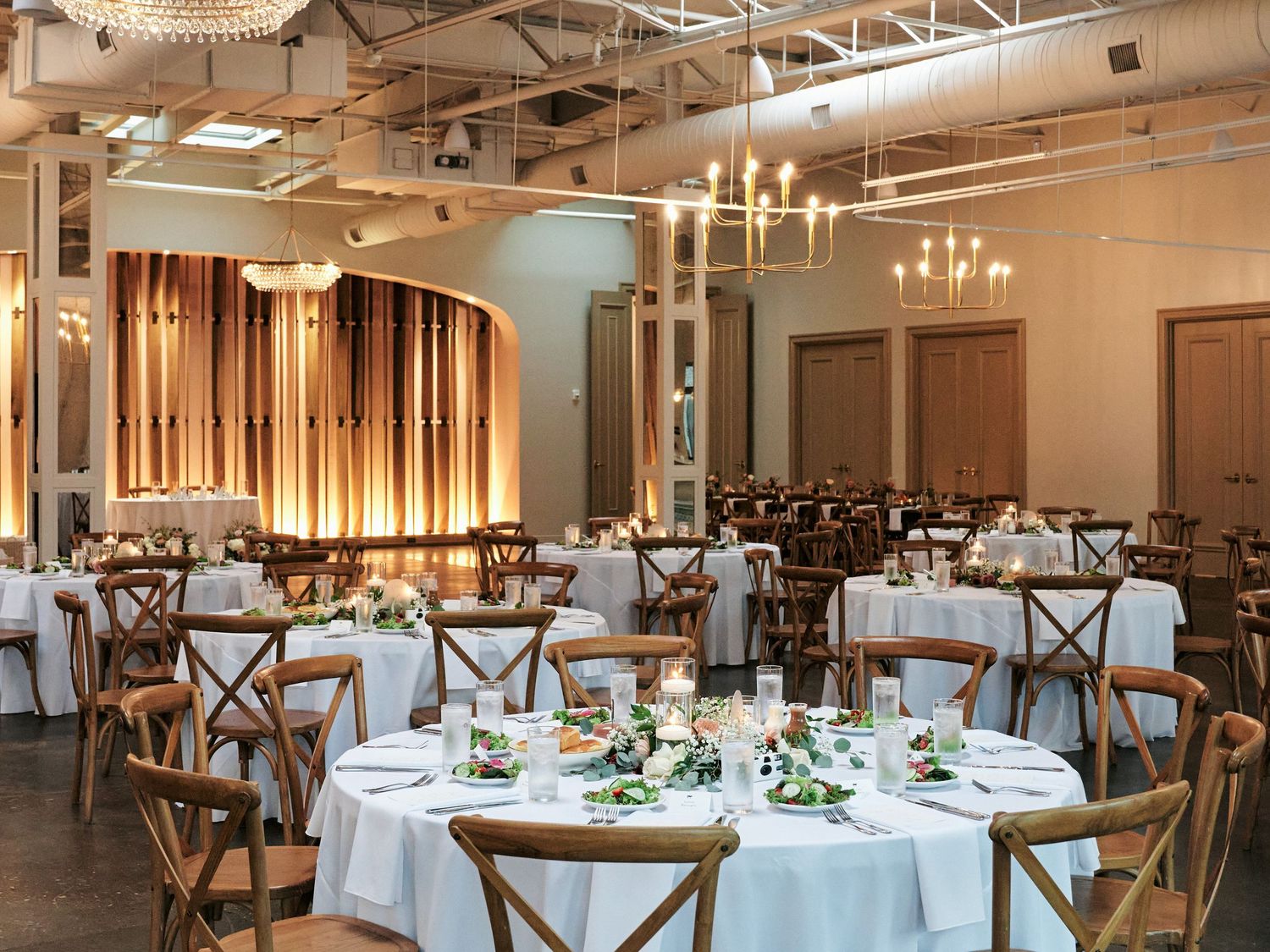 Elegant wedding reception hall with round tables, white linens, greenery centerpieces, wood cross-back chairs, and warm chandelier lighting.