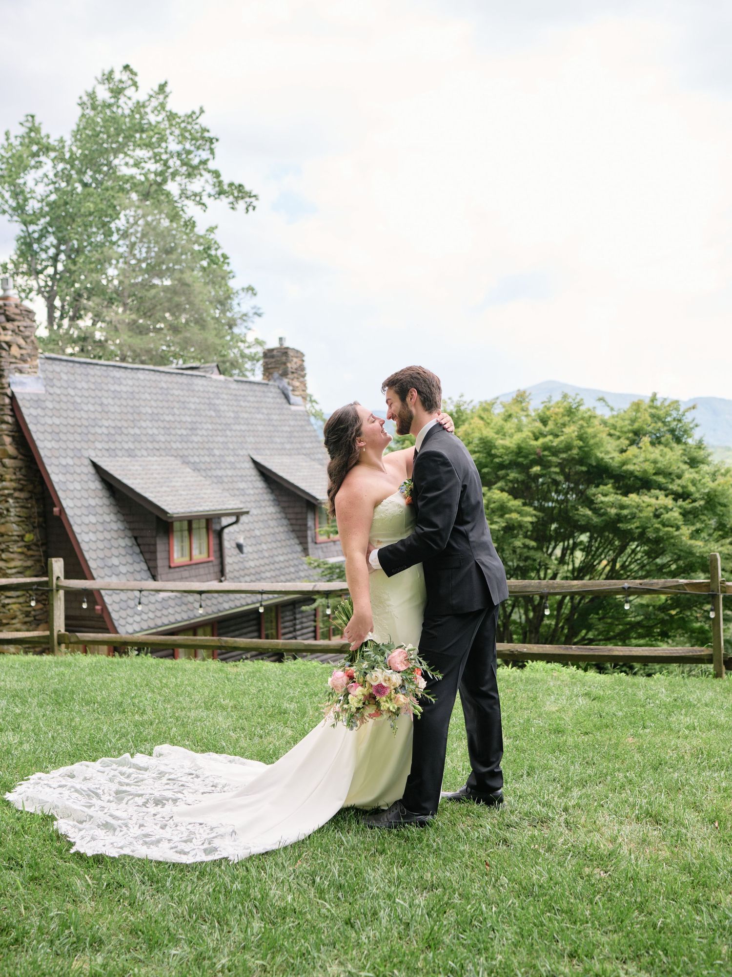 Bride in green gown and groom in black tux embrace on lawn before rustic stone chapel with mountain views.