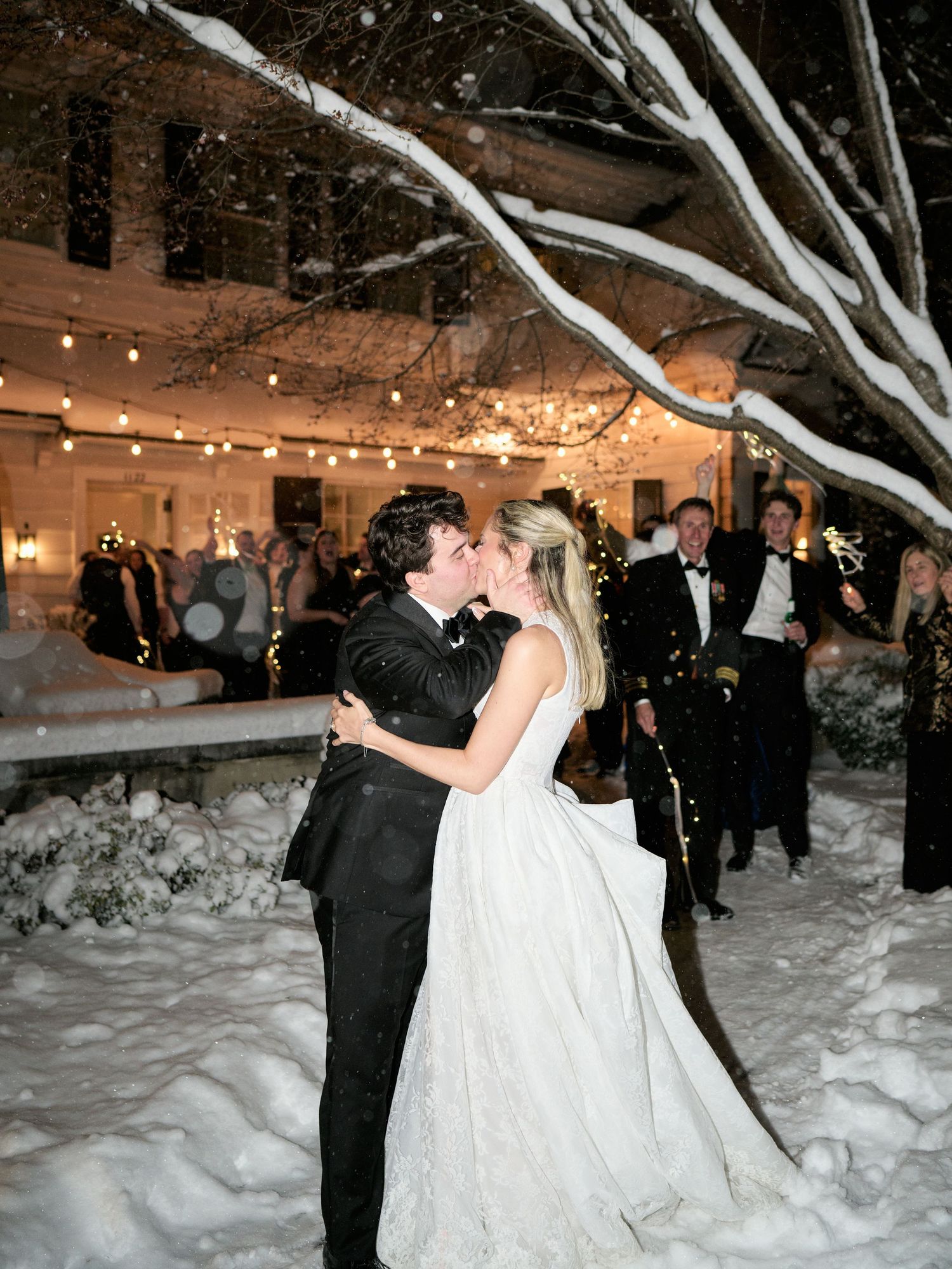Bride and groom share a romantic kiss in a snowy outdoor winter wedding setting with string lights and guests in formal attire.