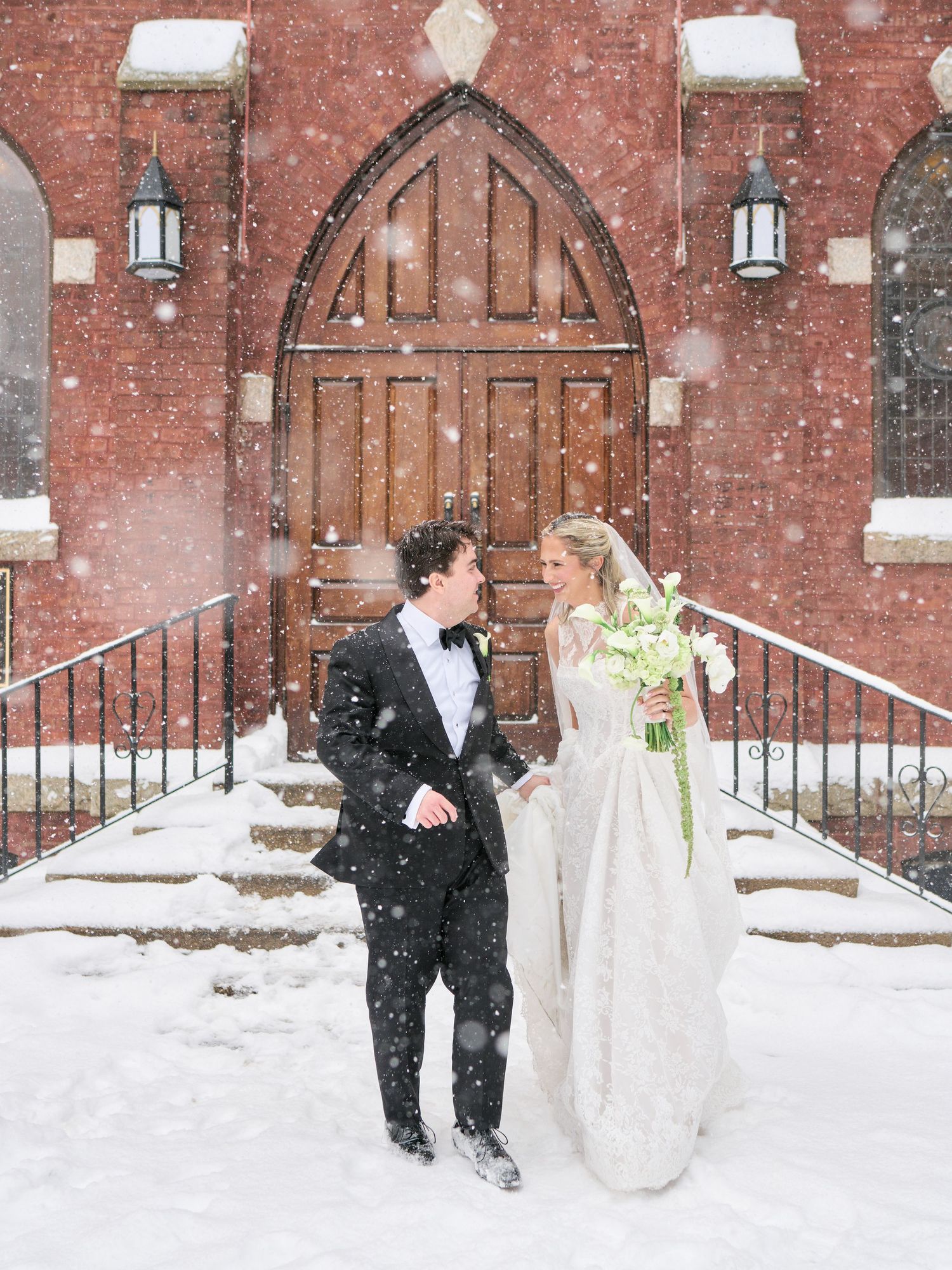 Bride and groom share a romantic moment in falling snow outside a brick church with wooden arched doors.