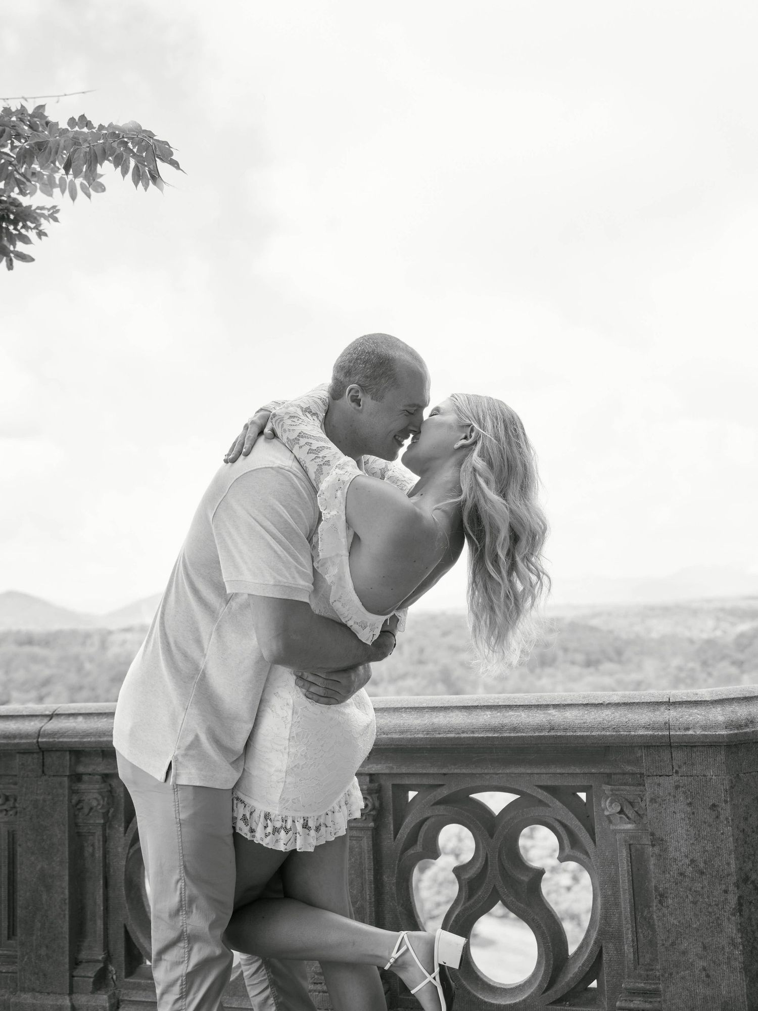 A romantic black and white photo of a couple sharing an intimate moment on a balcony with decorative stone railing.