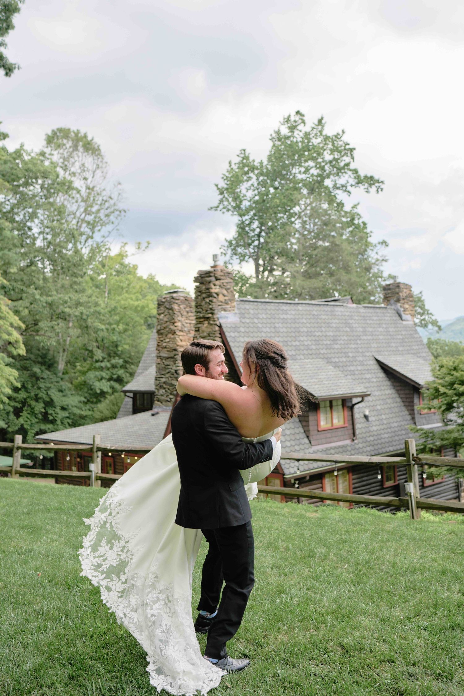 Groom lifts bride in white lace wedding gown outdoors on green lawn beside rustic stone and wood venue.
