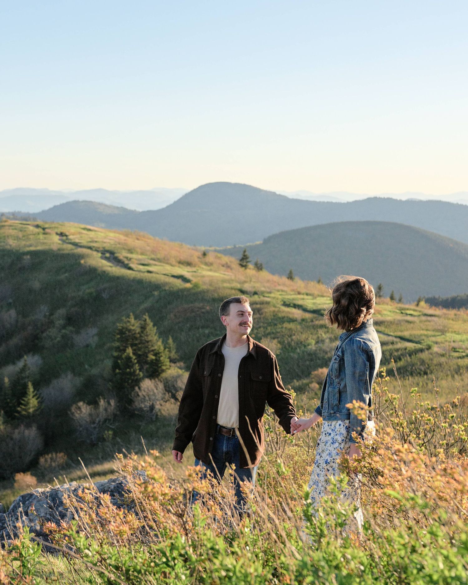 Couple holding hands on a scenic mountain overlook with rolling green hills and blue sky in the background.