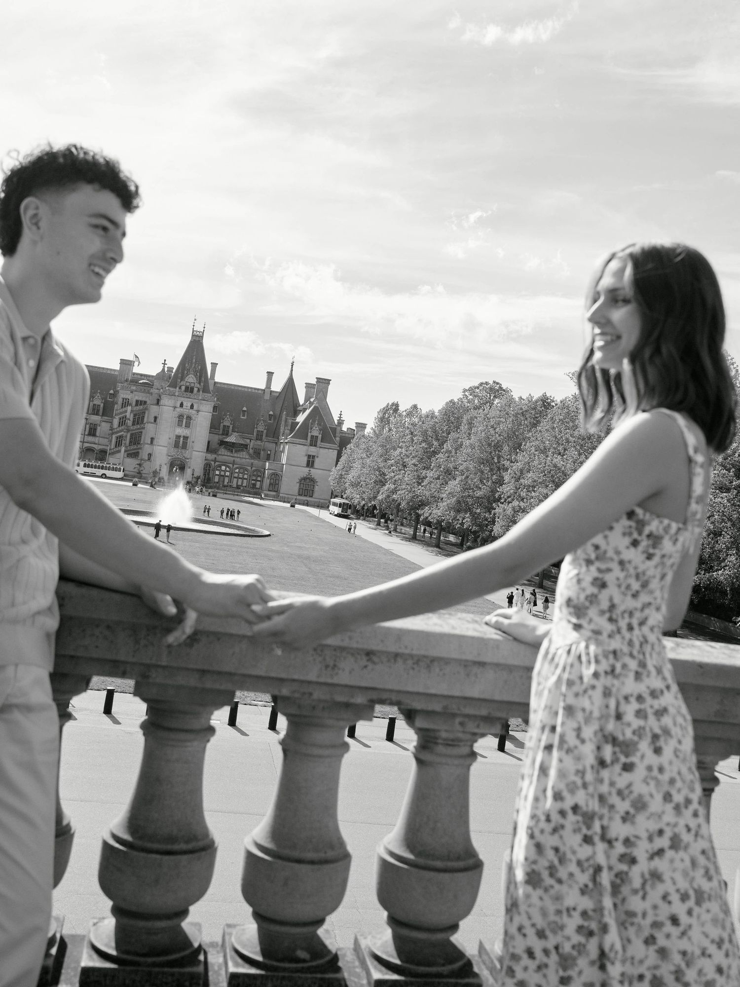 Black and white photo of a couple holding hands on a balustrade with a grand estate in the background.