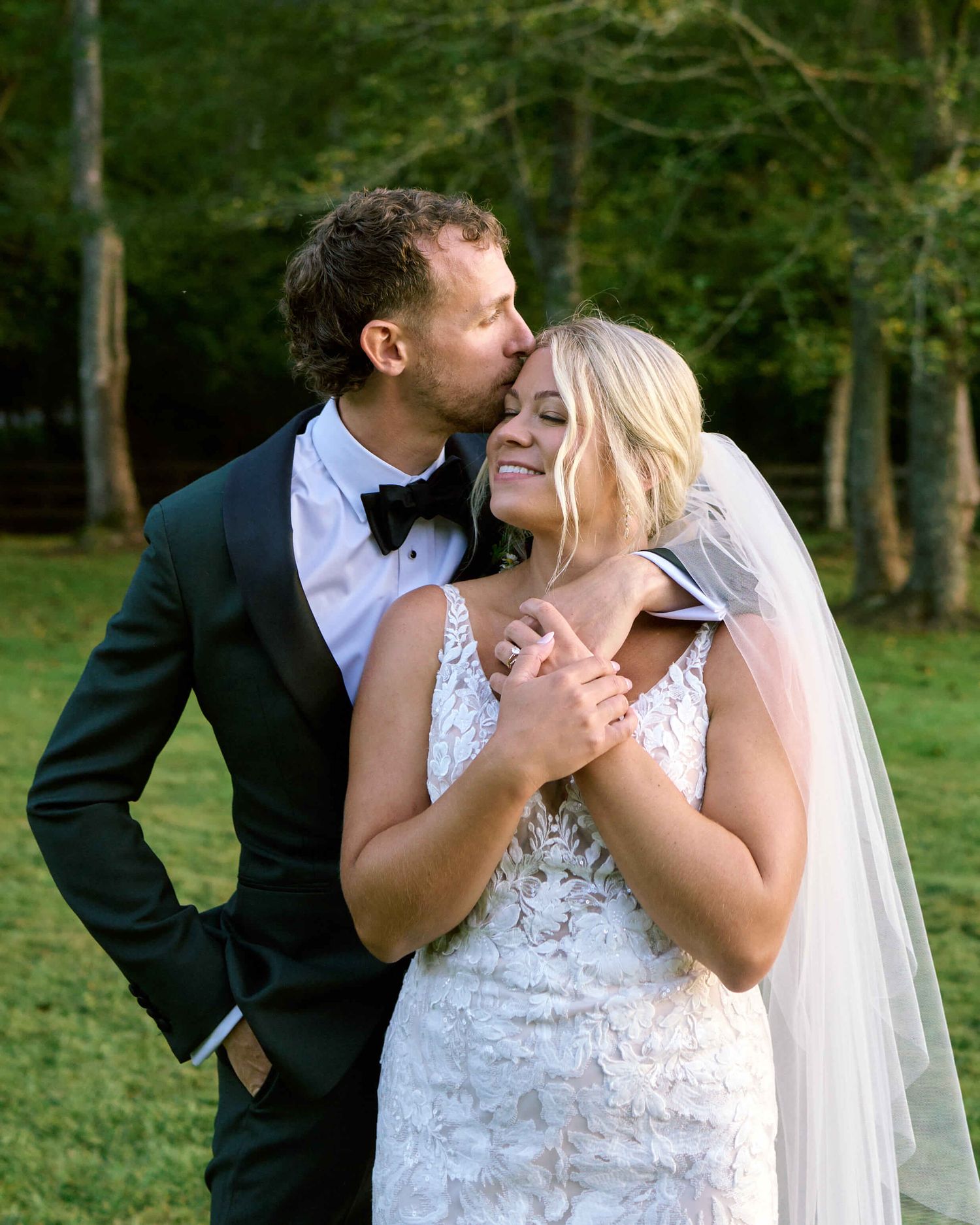 A couple shares a romantic kiss in a green outdoor setting during their wedding photoshoot.