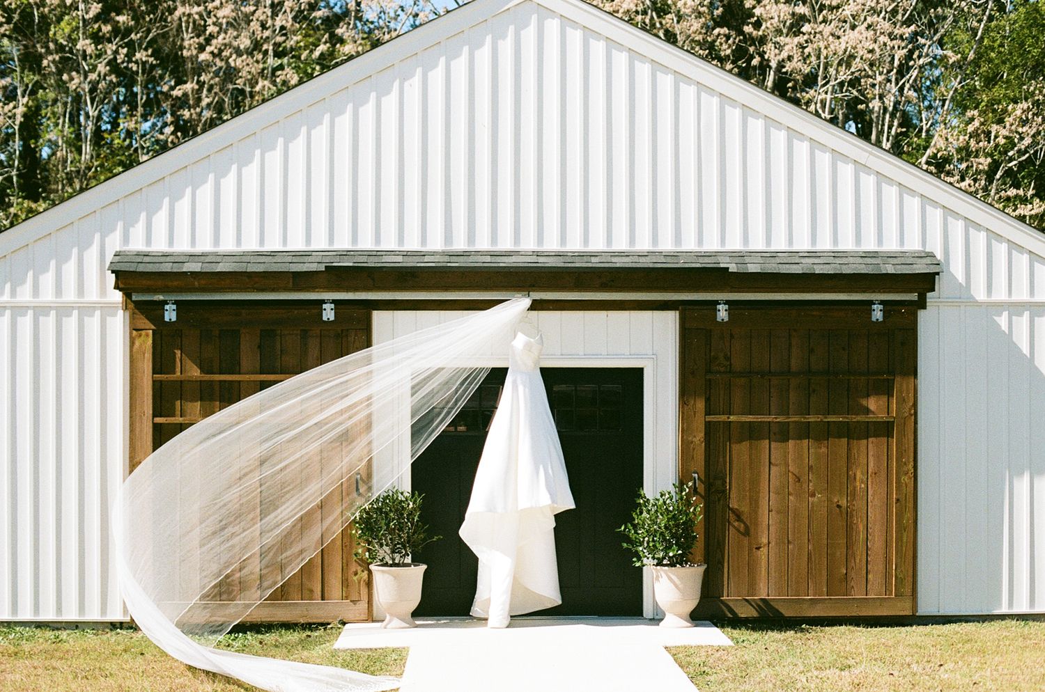 White wedding dresses and veils float ethereally in a rustic barn setting with wooden doors and white walls.