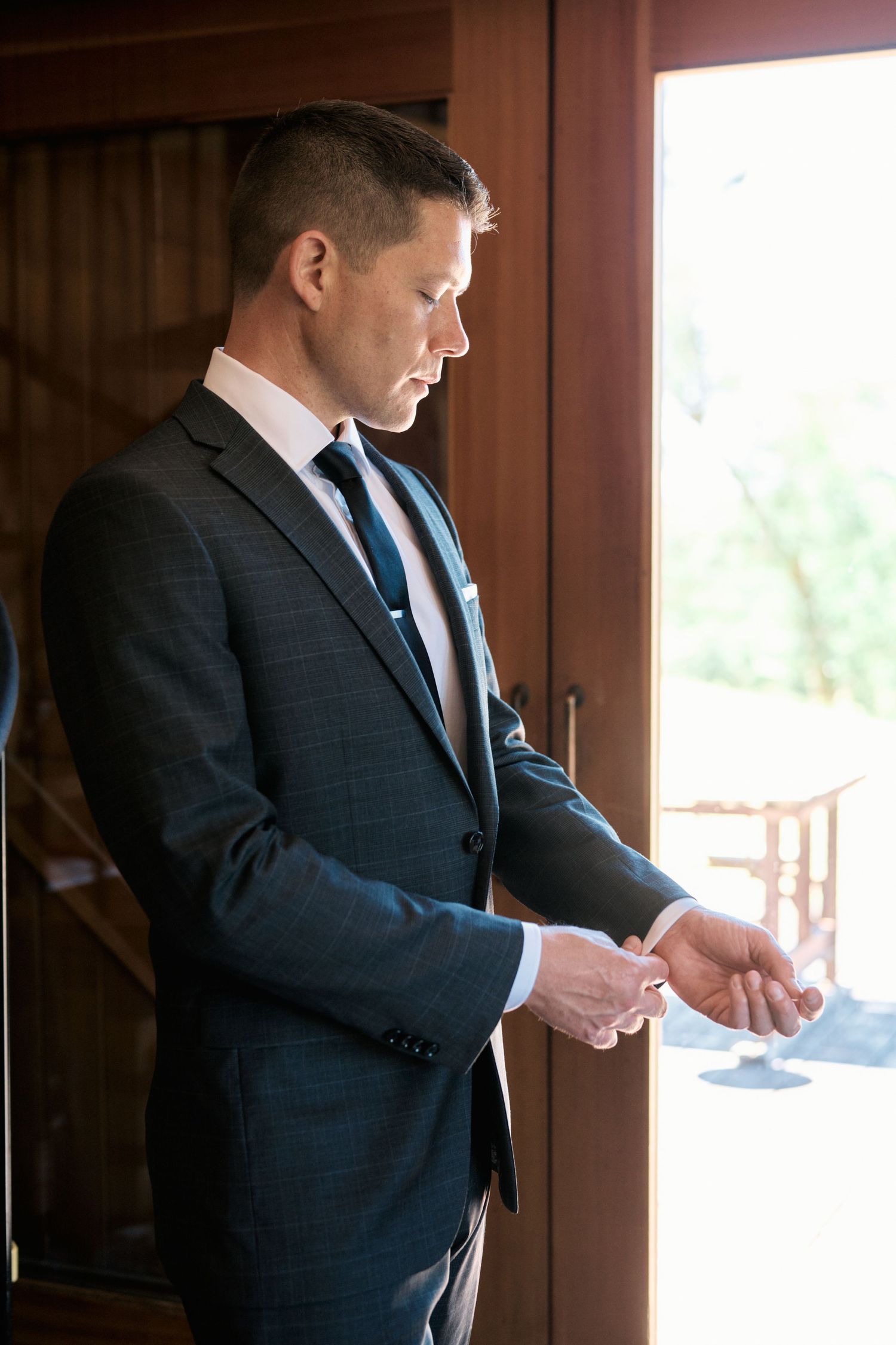 Person in dark suit adjusting cufflinks while standing by a window.
