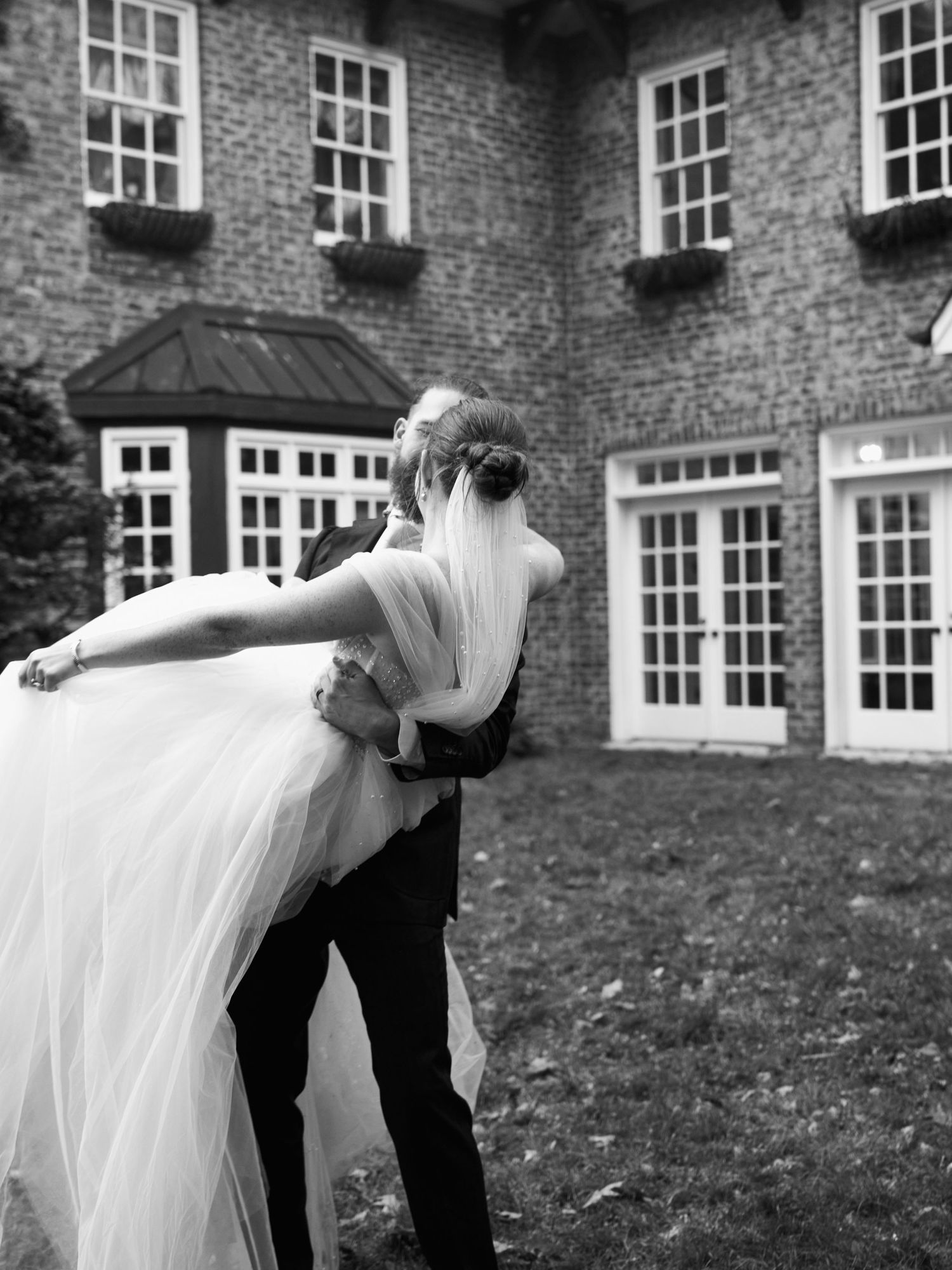 A dramatic black and white photo of newlyweds posing outside a brick mansion.