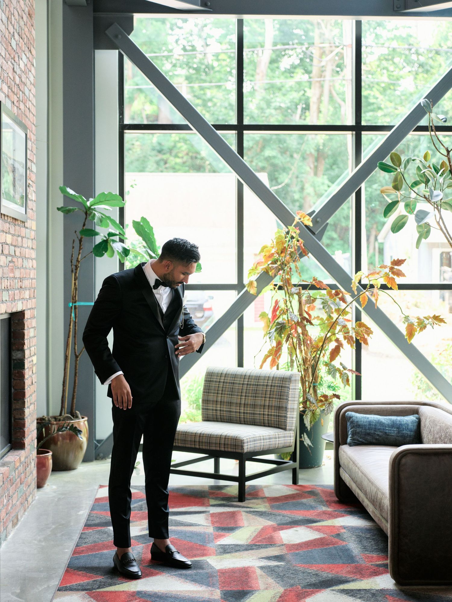 Well-dressed person in black suit stands in modern office space with geometric windows, plants and patterned rug.