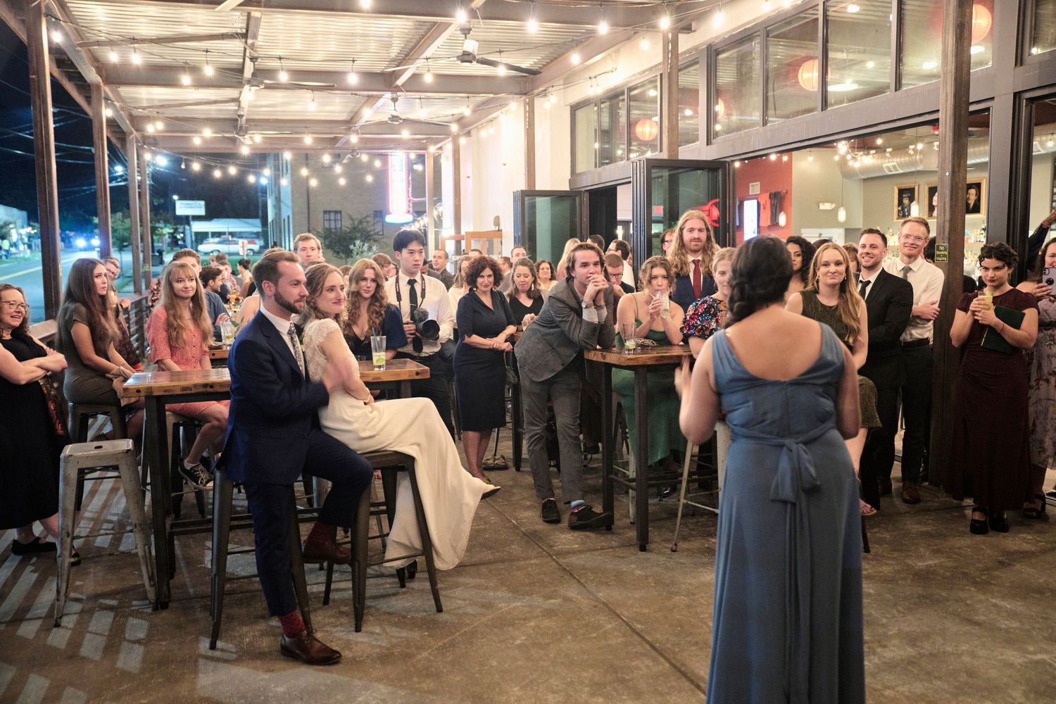 Wedding guests gather in an industrial-style venue with exposed brick walls and hanging string lights.