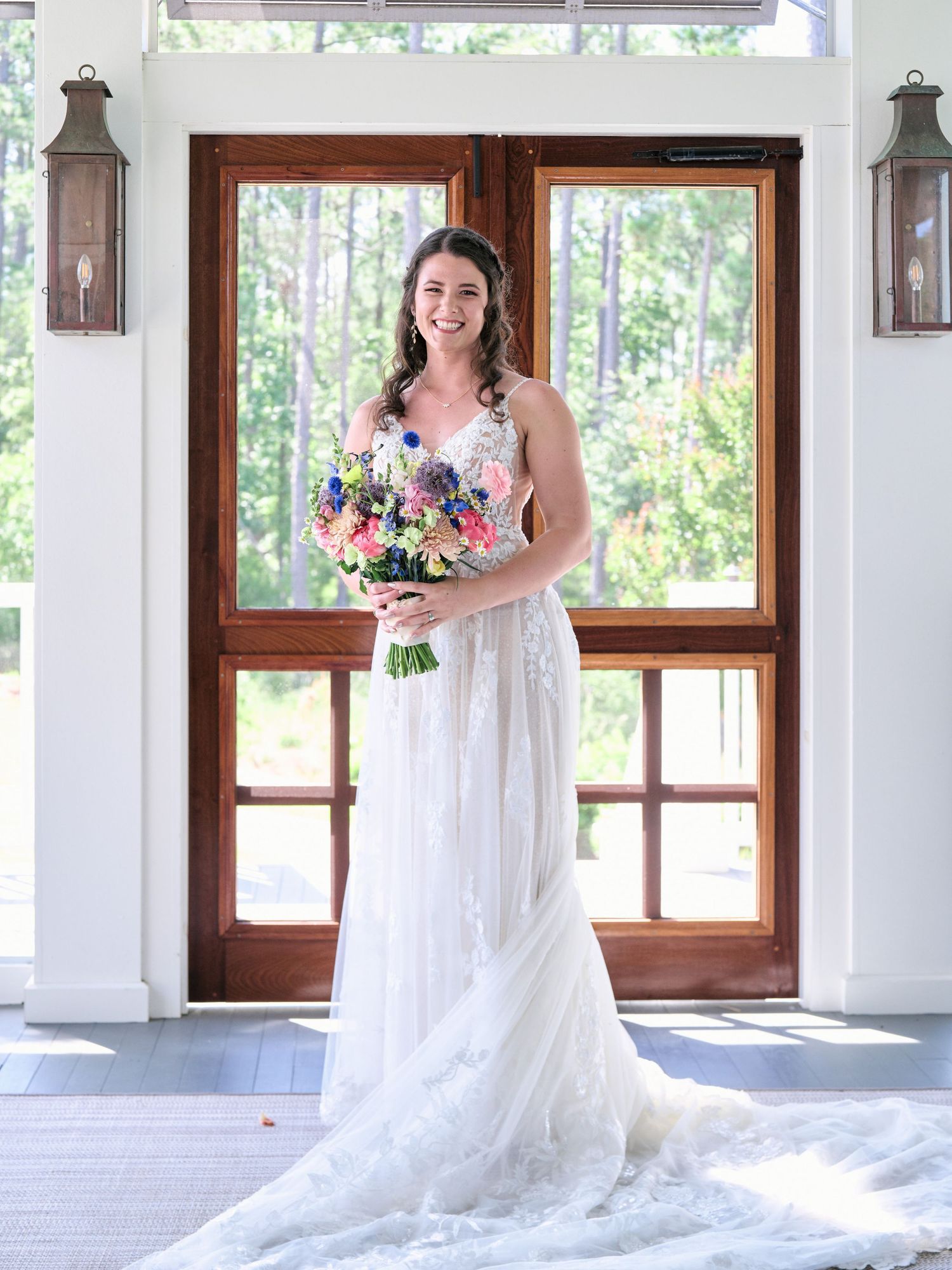 White lace wedding dress with a flowing train and colorful summer bouquet photographed in front of wooden French doors.