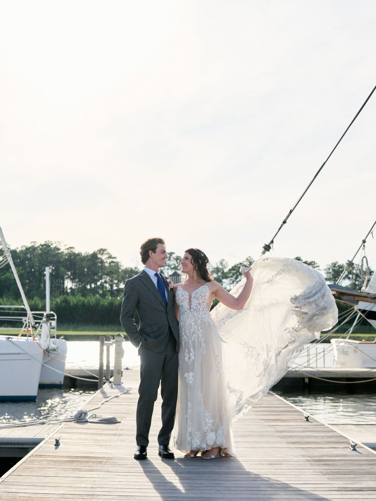 Elegant newlyweds share a romantic moment on a marina dock with sailboats and water in the background.