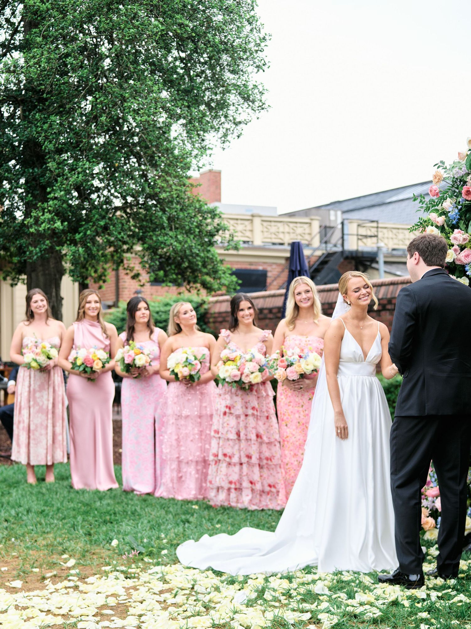 Bridal party lined up outdoors with bridesmaids wearing pink dresses against a brick wall backdrop.