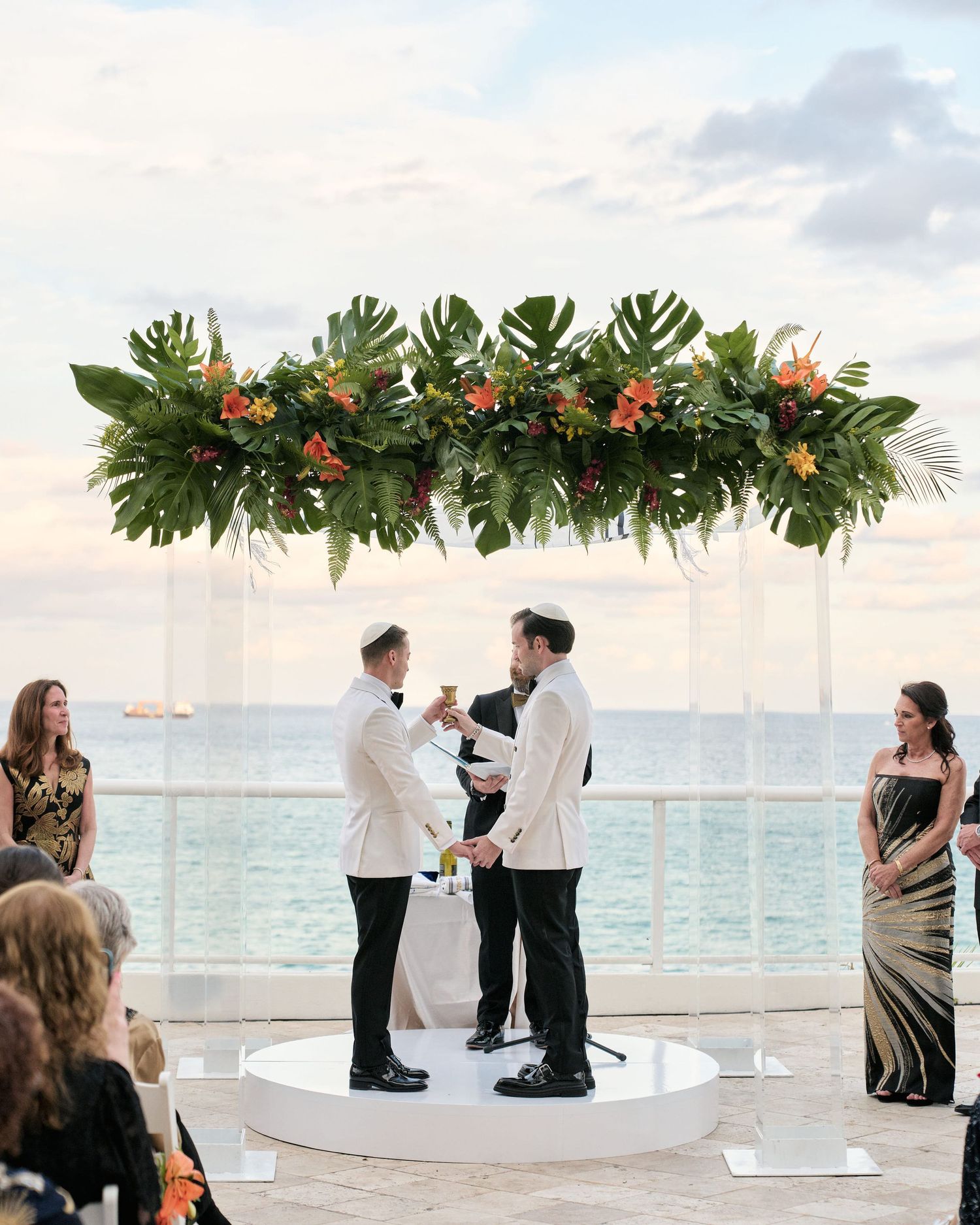 Beautiful beach wedding ceremony under a tropical floral arch with ocean view and sunset sky backdrop.