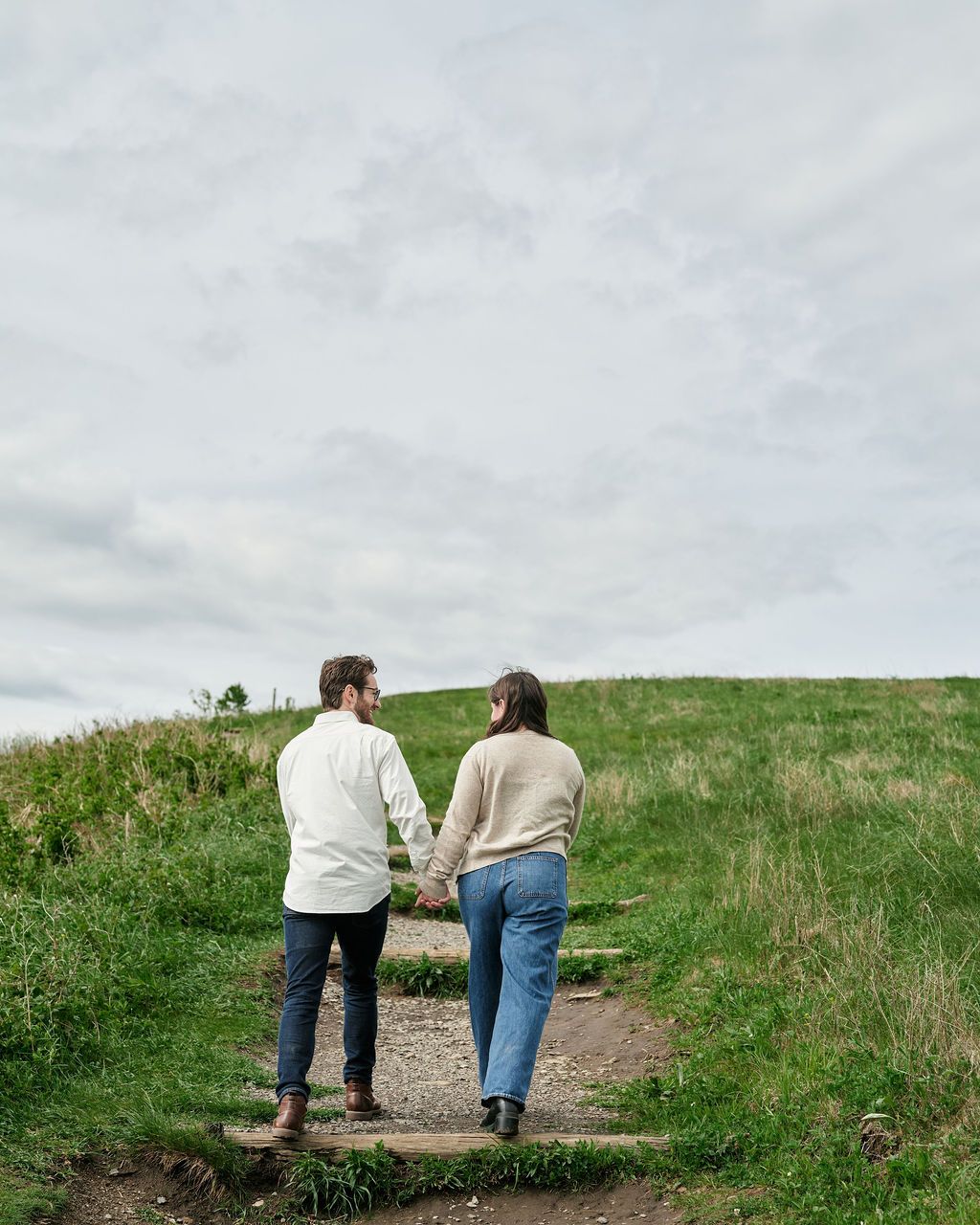 A couple walks hand in hand along a scenic grassy hilltop path with cloudy skies overhead.