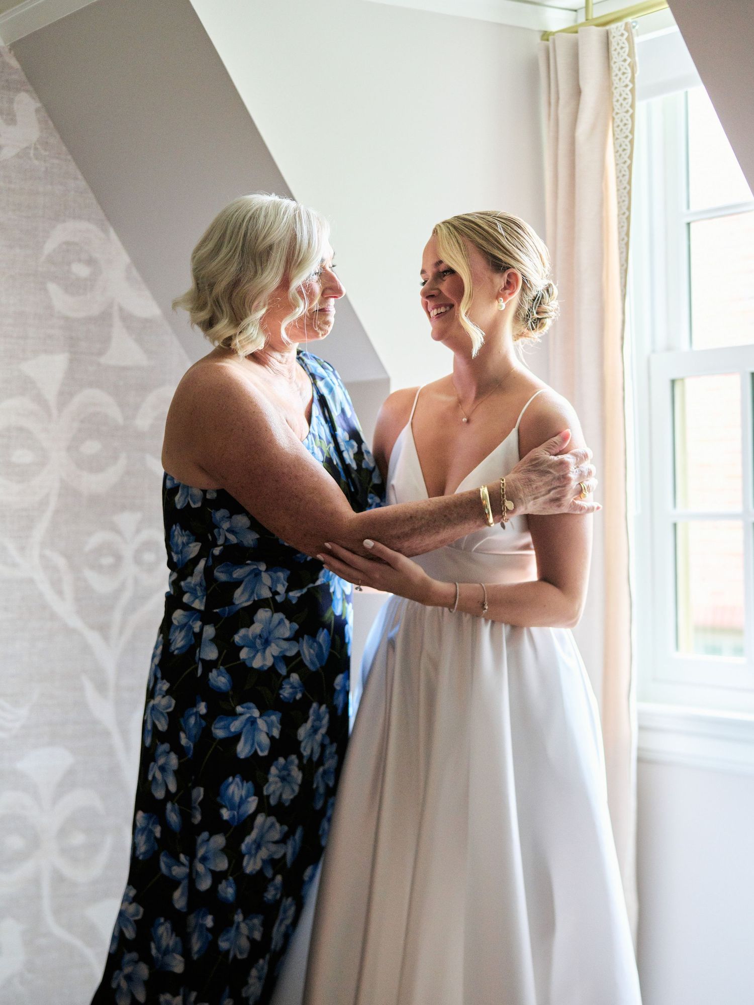 Two women share an emotional moment in a sunlit room, one wearing a floral dress and one in a white wedding gown.