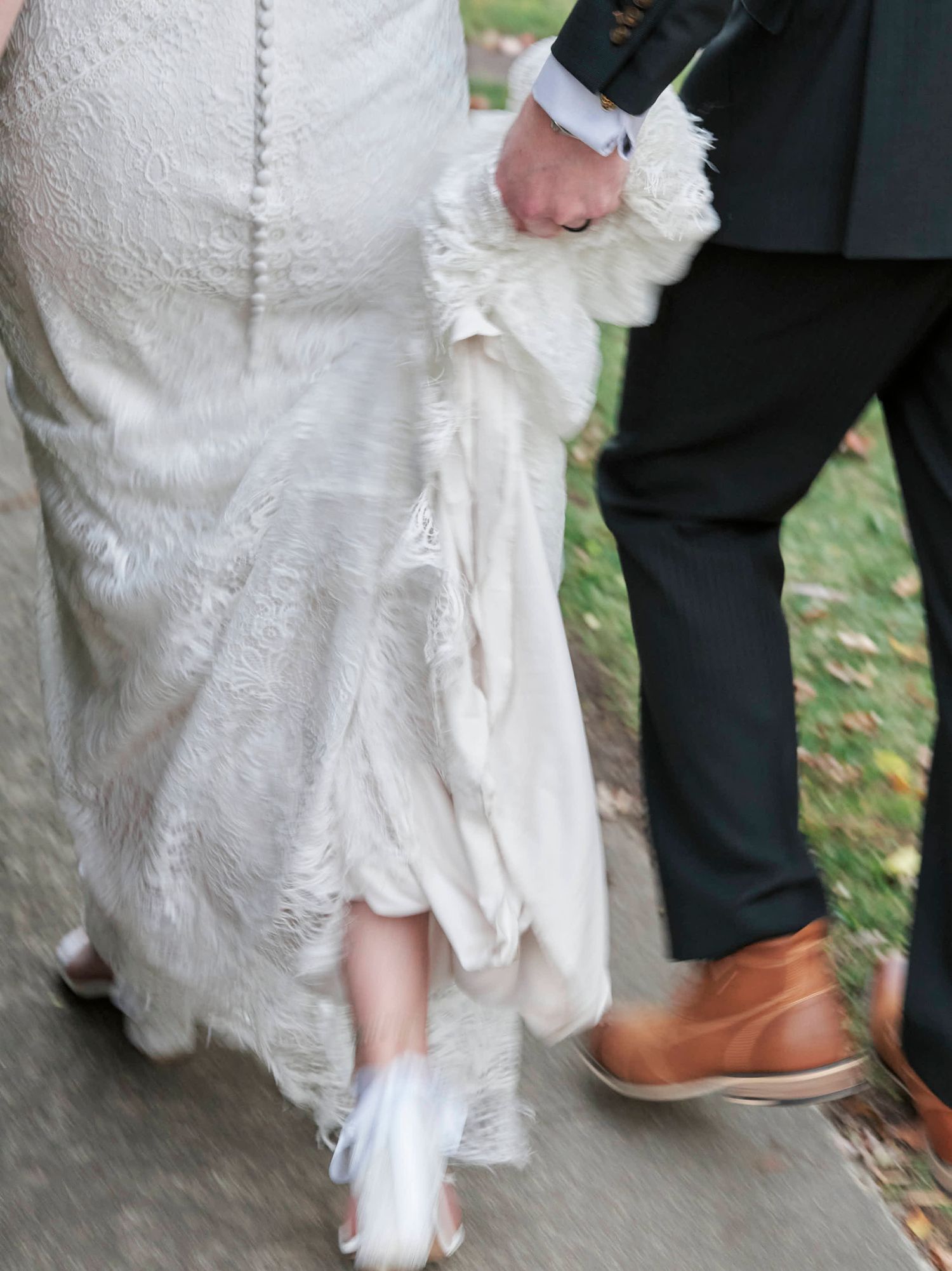 Close-up of white wedding dress hem and brown leather shoes on a stone pathway.