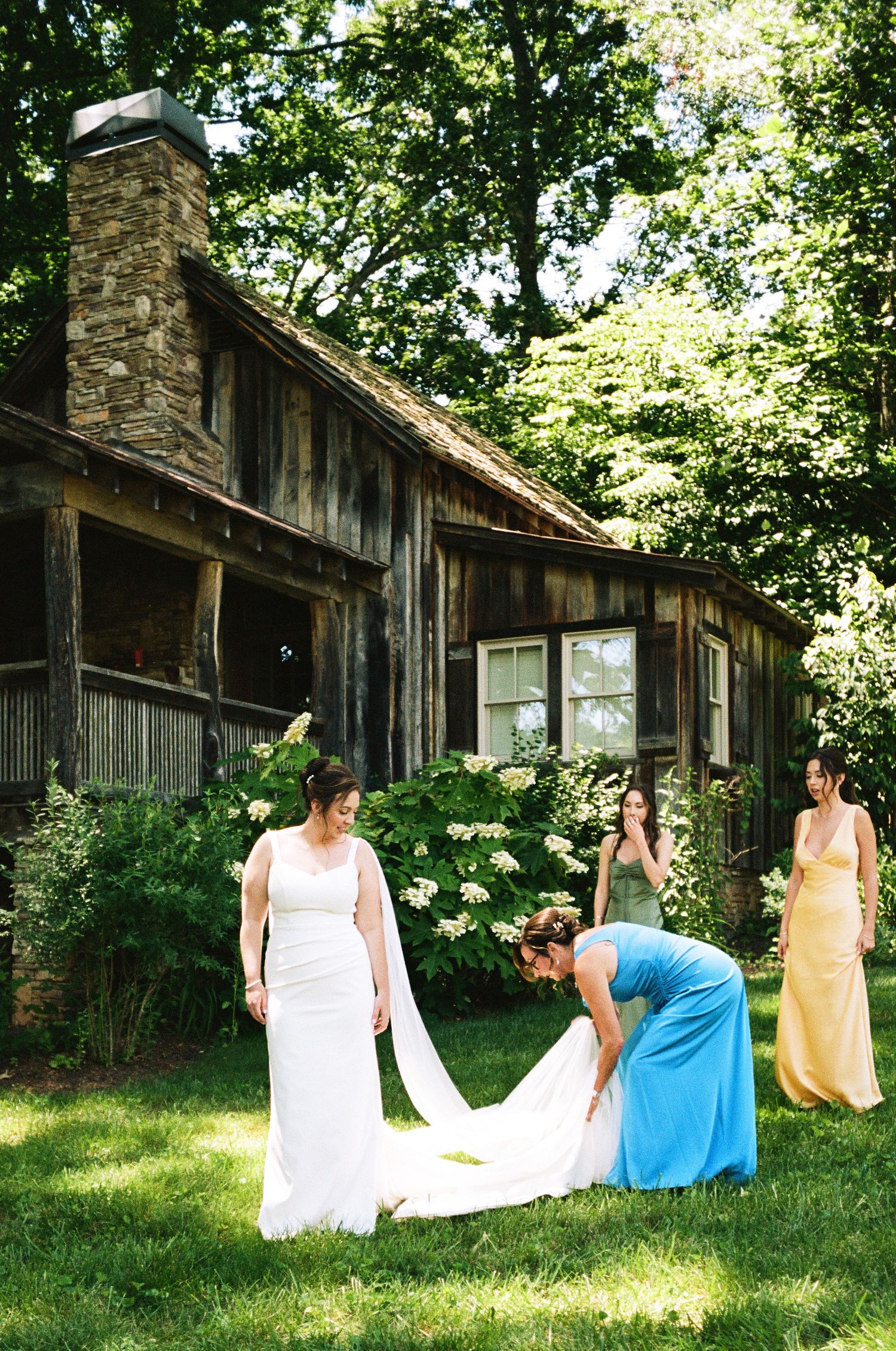 Bridesmaids in colorful gowns help bride with white wedding dress train on green lawn outside rustic stone cabin.