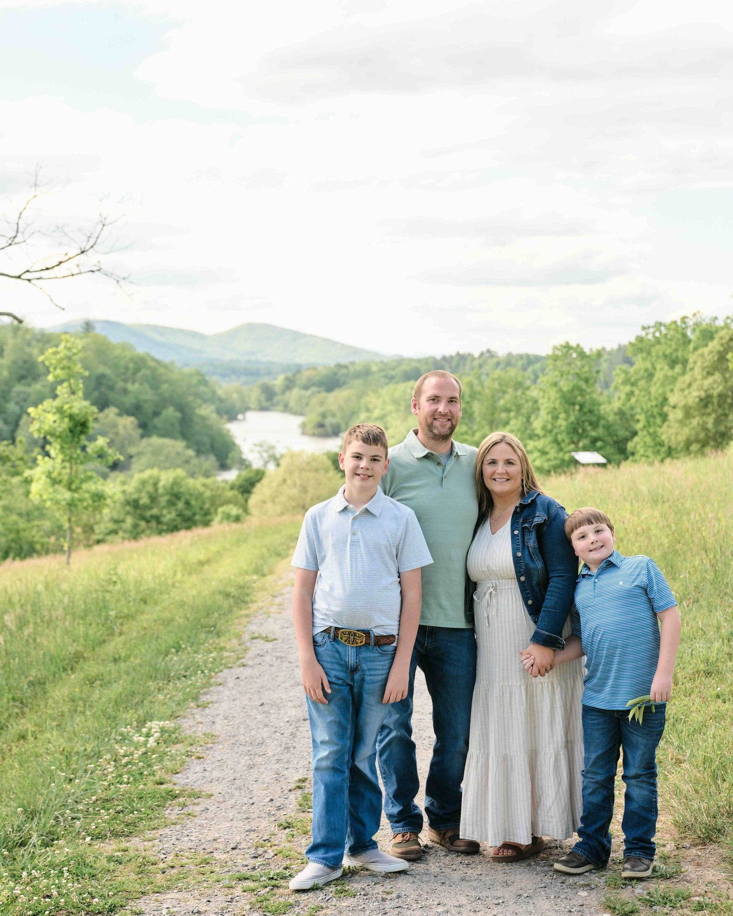 Family of four smiling on a scenic gravel path with rolling green hills and a river in the background.