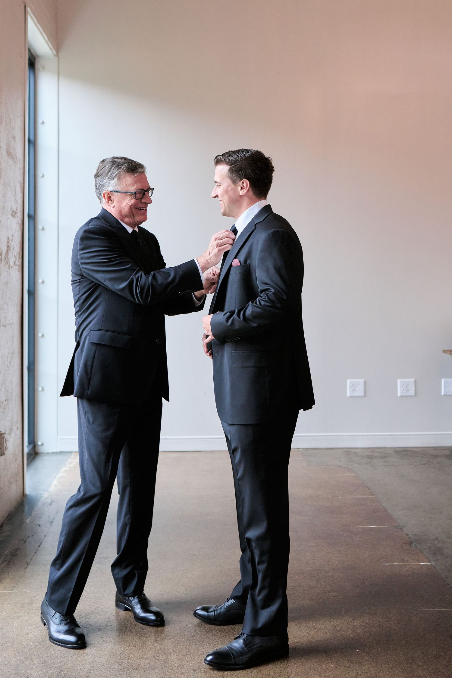 An older man in a black suit adjusts the tie of a younger man in a dark suit in a bright modern room.