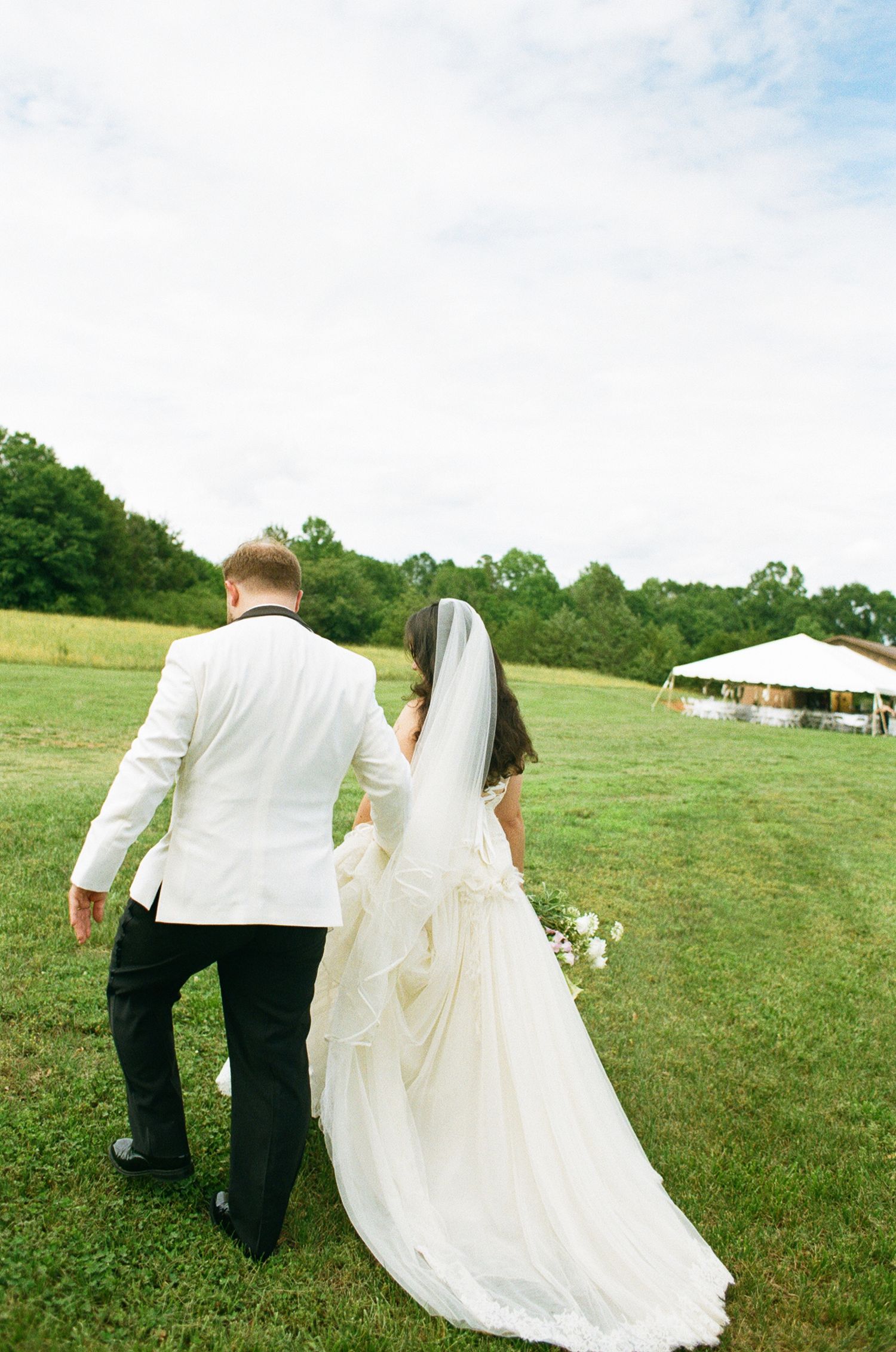 Bride and groom walking hand in hand across green field, bride in white gown with veil, groom in white jacket.