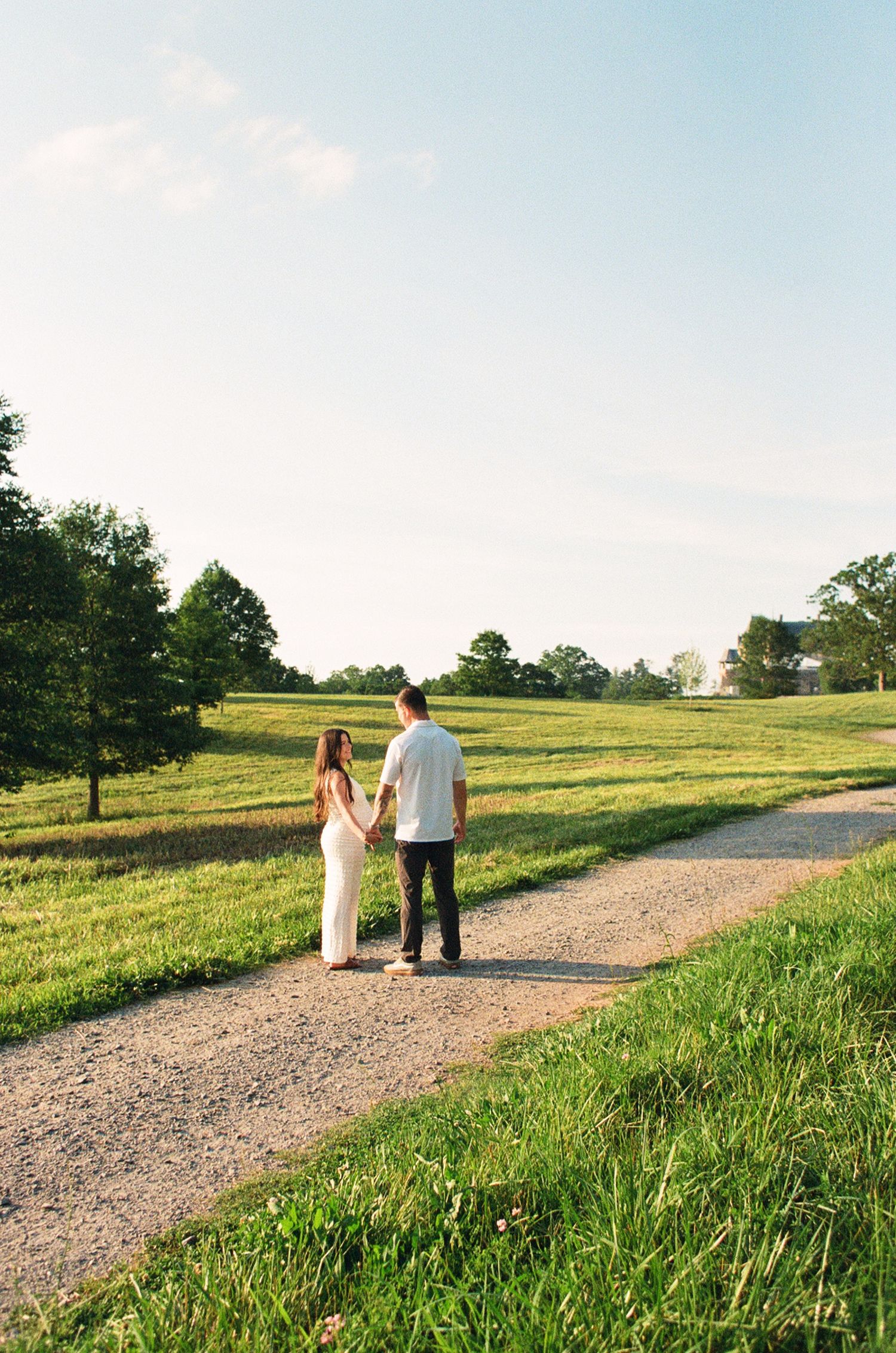 A couple holds hands while standing on a gravel path surrounded by lush green fields and trees on a sunny day.
