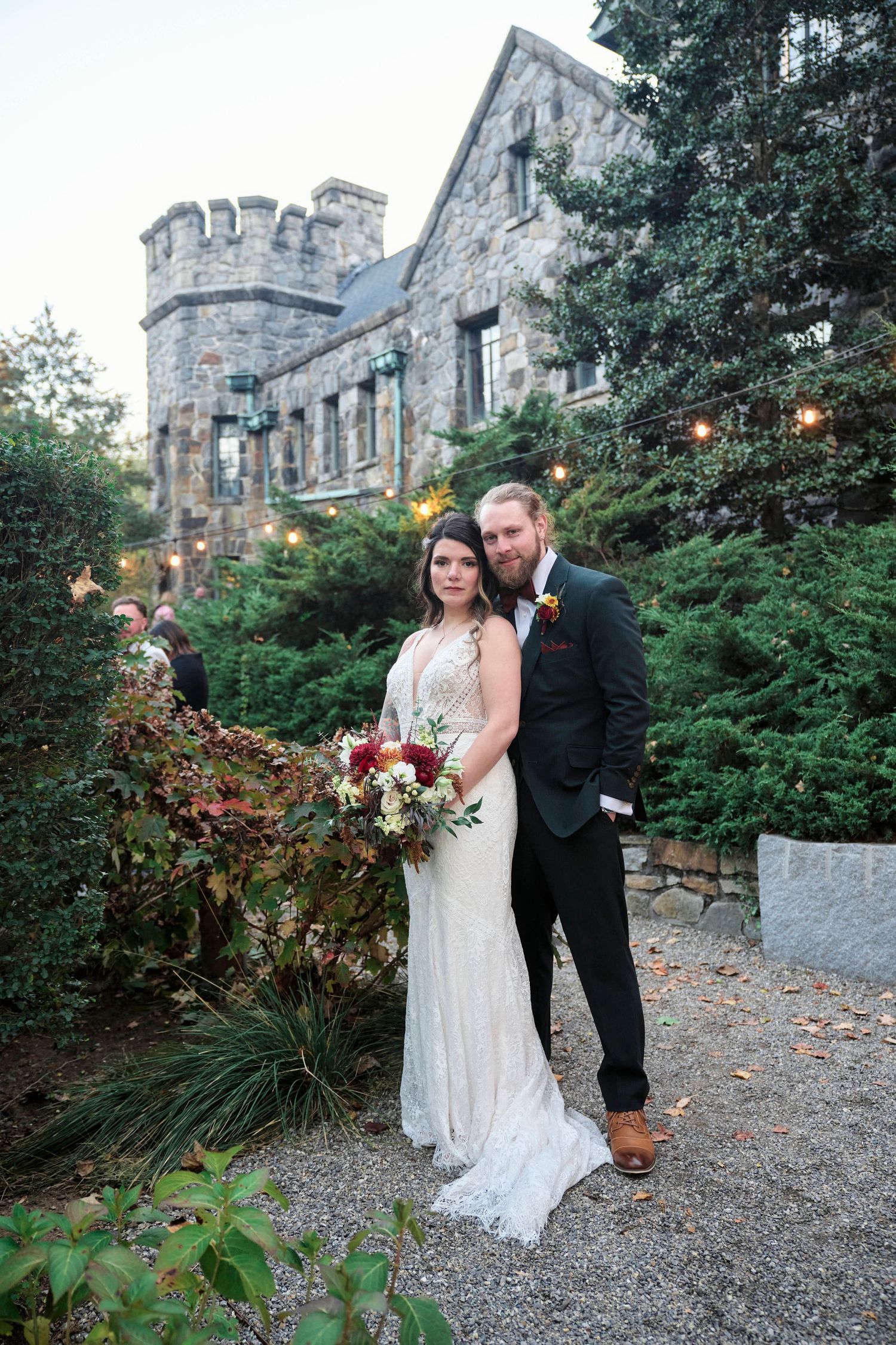 Wedding couple poses in front of a stone castle with turret and lush landscaping.
