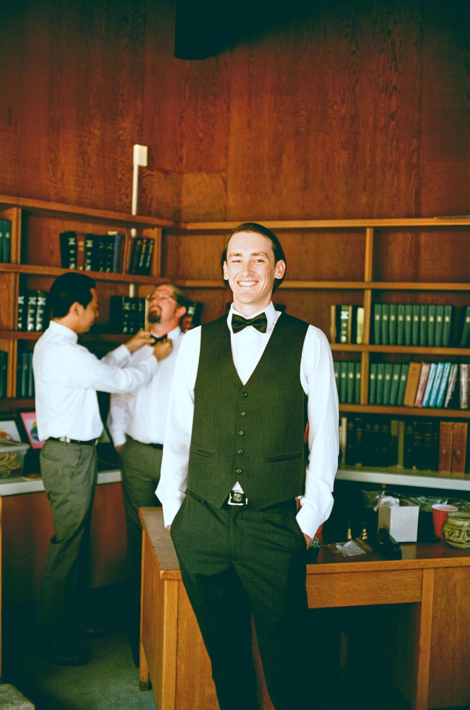 Wedding party member adjusting his bowtie in a room with wooden bookshelves.