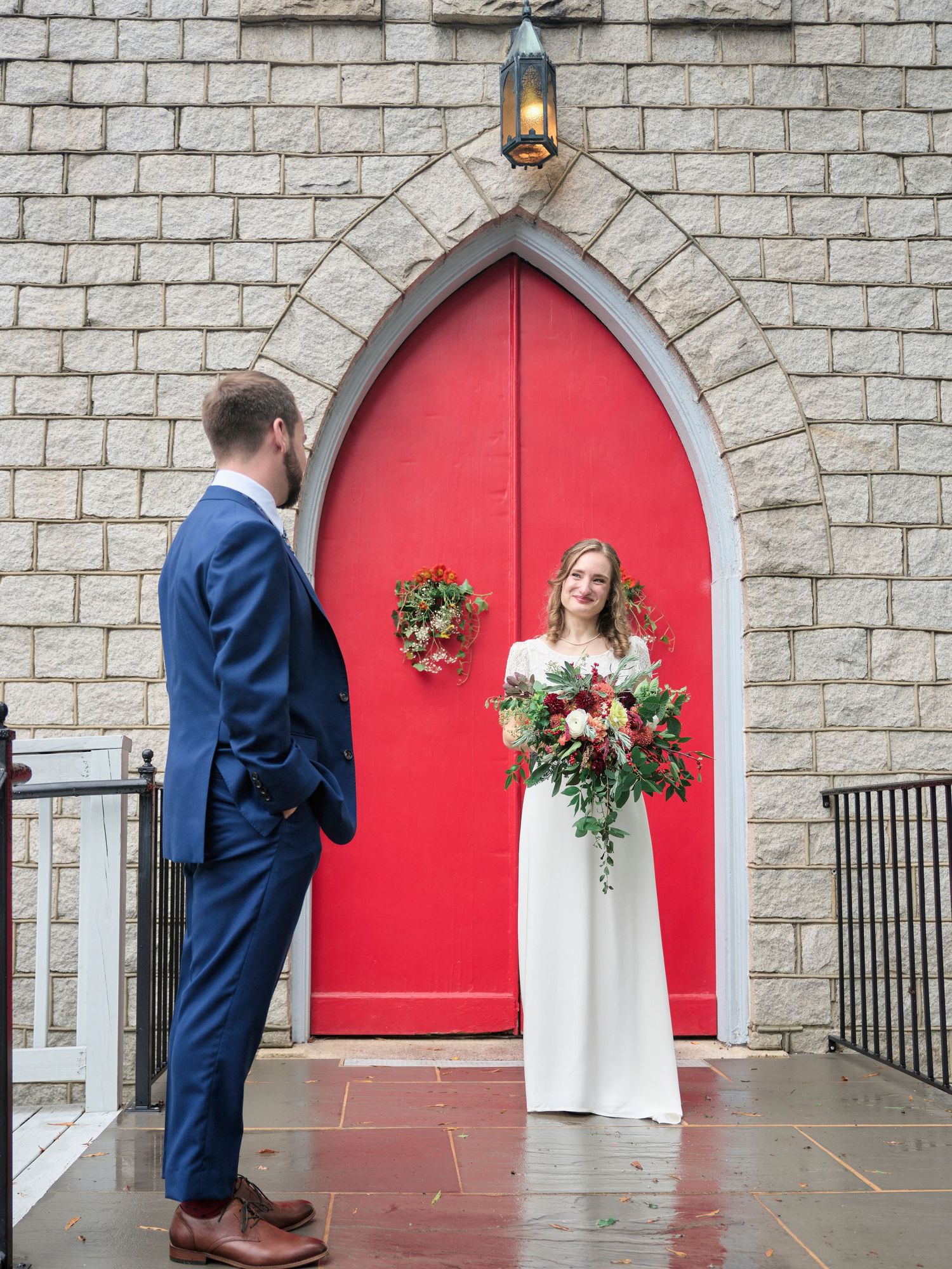 A wedding couple poses in front of a striking red church door framed by stone architecture.