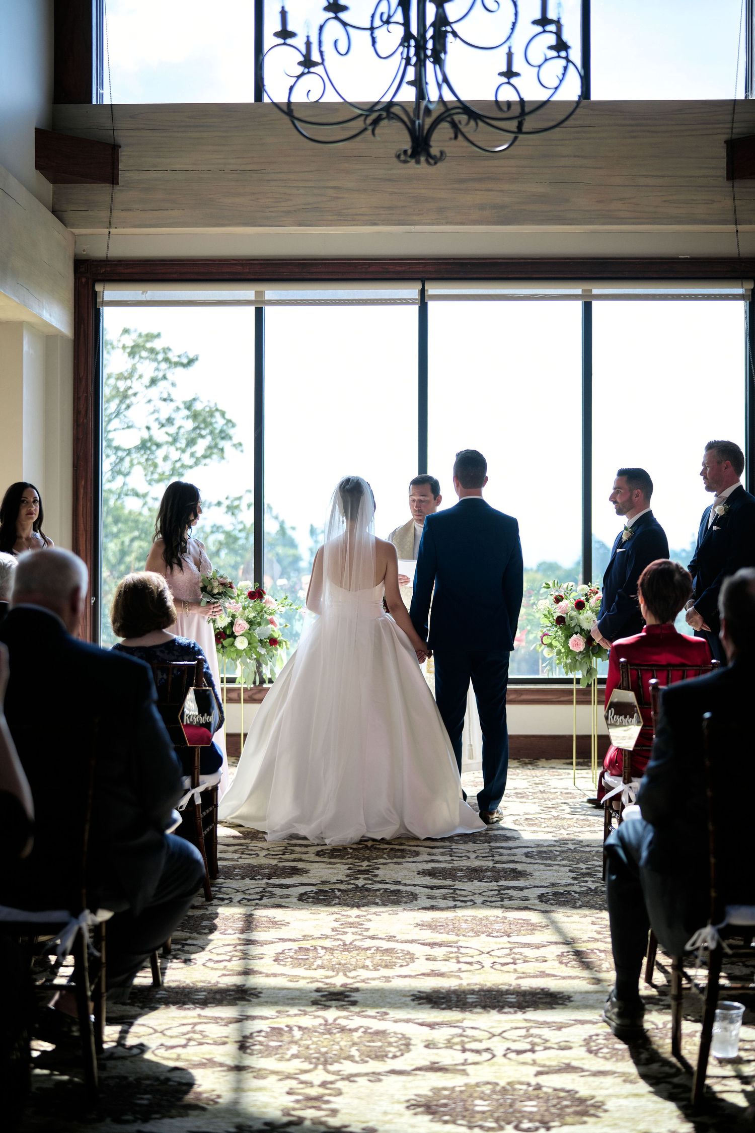 Wedding ceremony in progress with elegant chandelier and floor-to-ceiling windows.