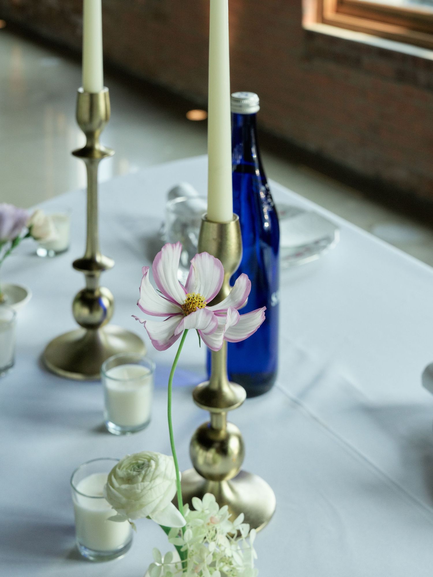 Elegant table setting with white candles in brass holders, blue bottle, and delicate cosmos flowers on crisp white tablecloth.