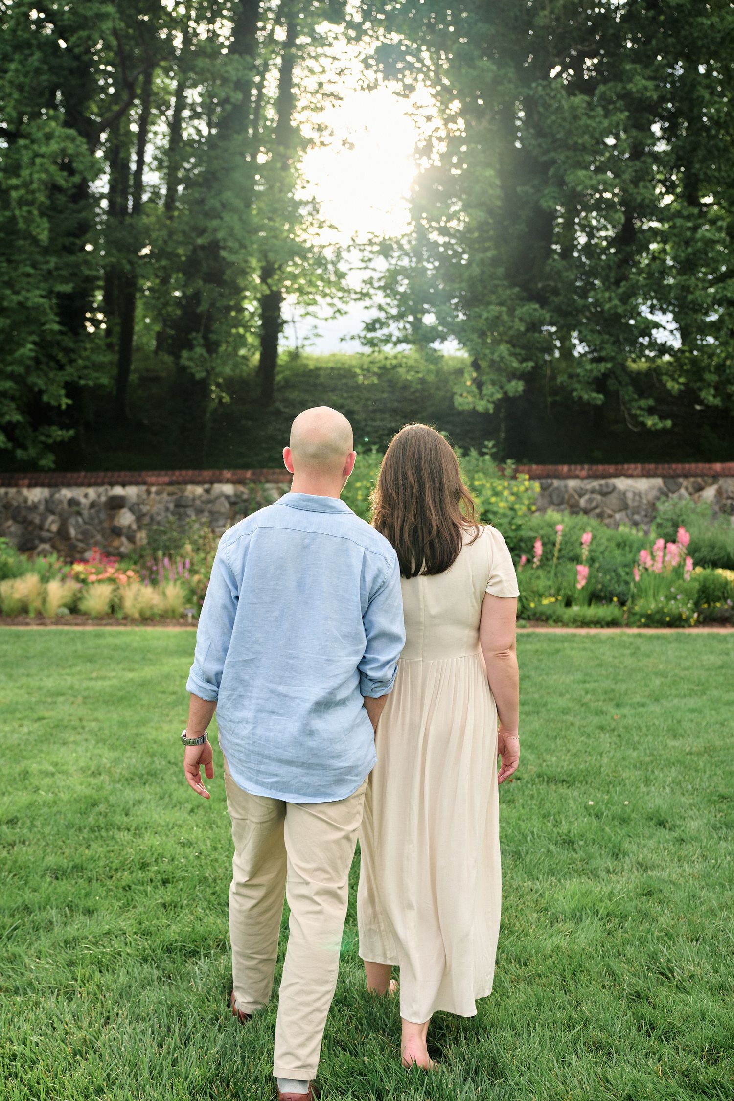 A couple stands together looking at a sunset through trees in a beautiful garden setting.