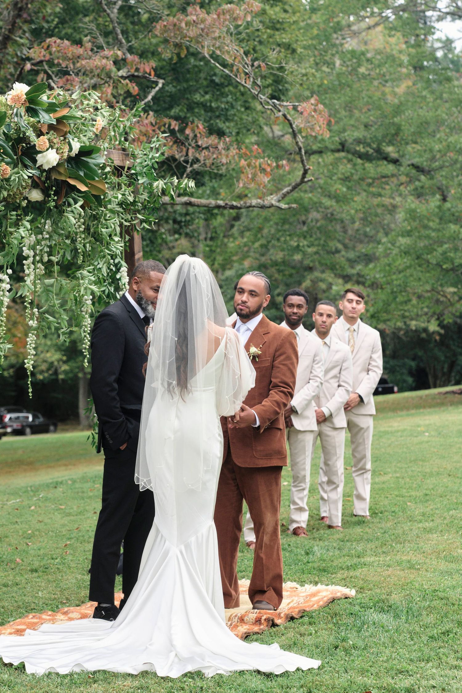 Wedding ceremony under floral arch with bridal party lined up on grassy lawn.