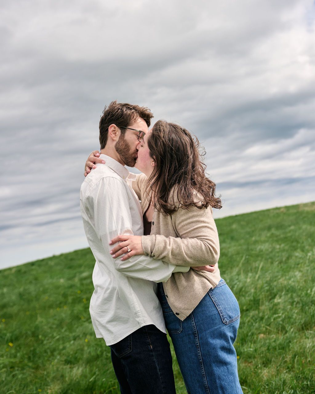 A romantic couple embraces and shares a kiss on a grassy hillside under dramatic cloudy skies.