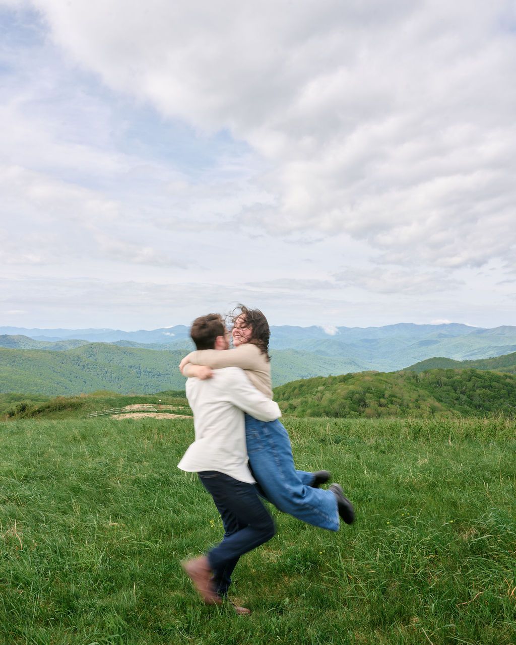 A couple embraces in a joyful spin on a grassy mountaintop meadow with rolling hills and cloudy skies in the background.
