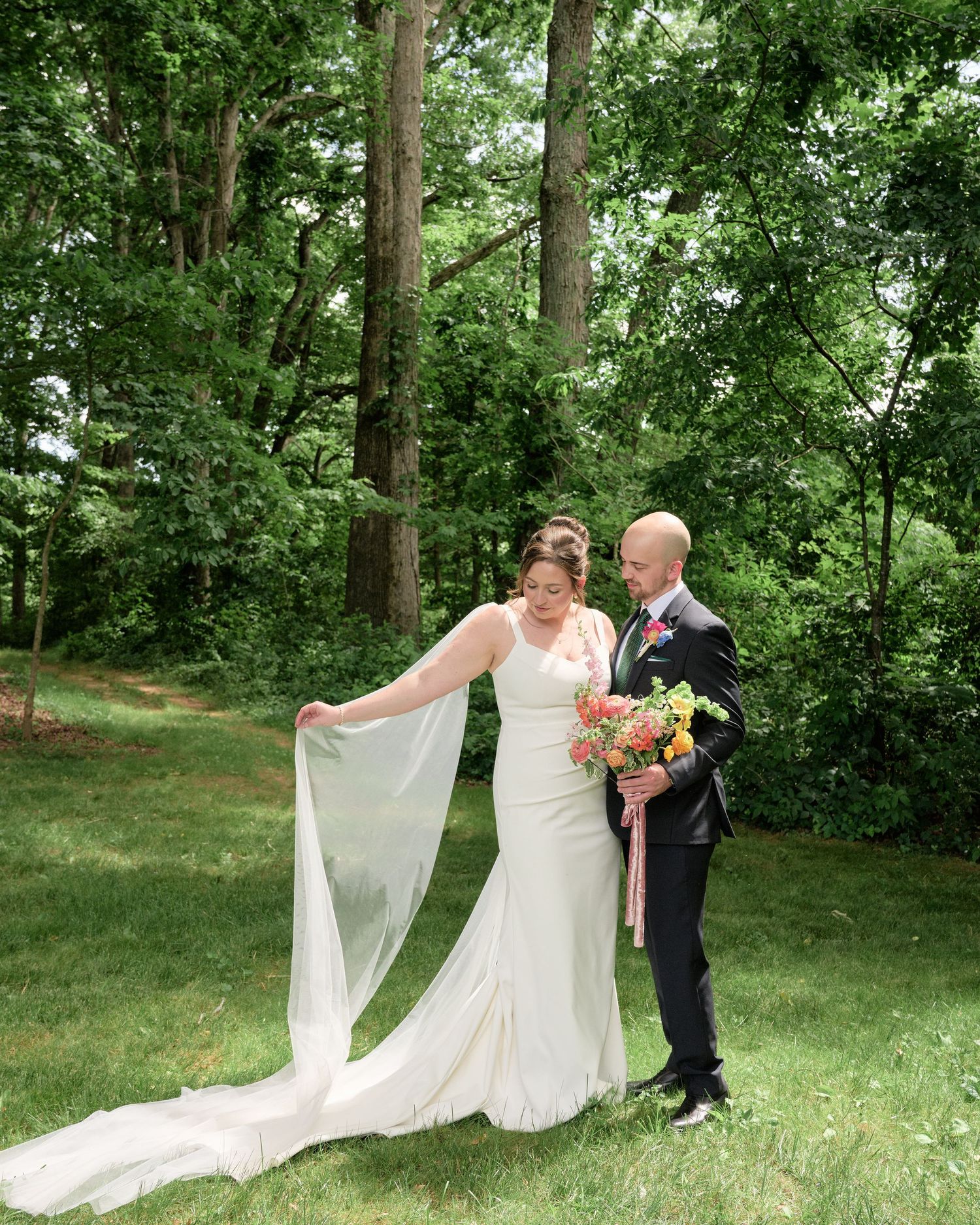 Newlyweds pose together on grassy lawn as bride's veil flows gracefully in outdoor setting.