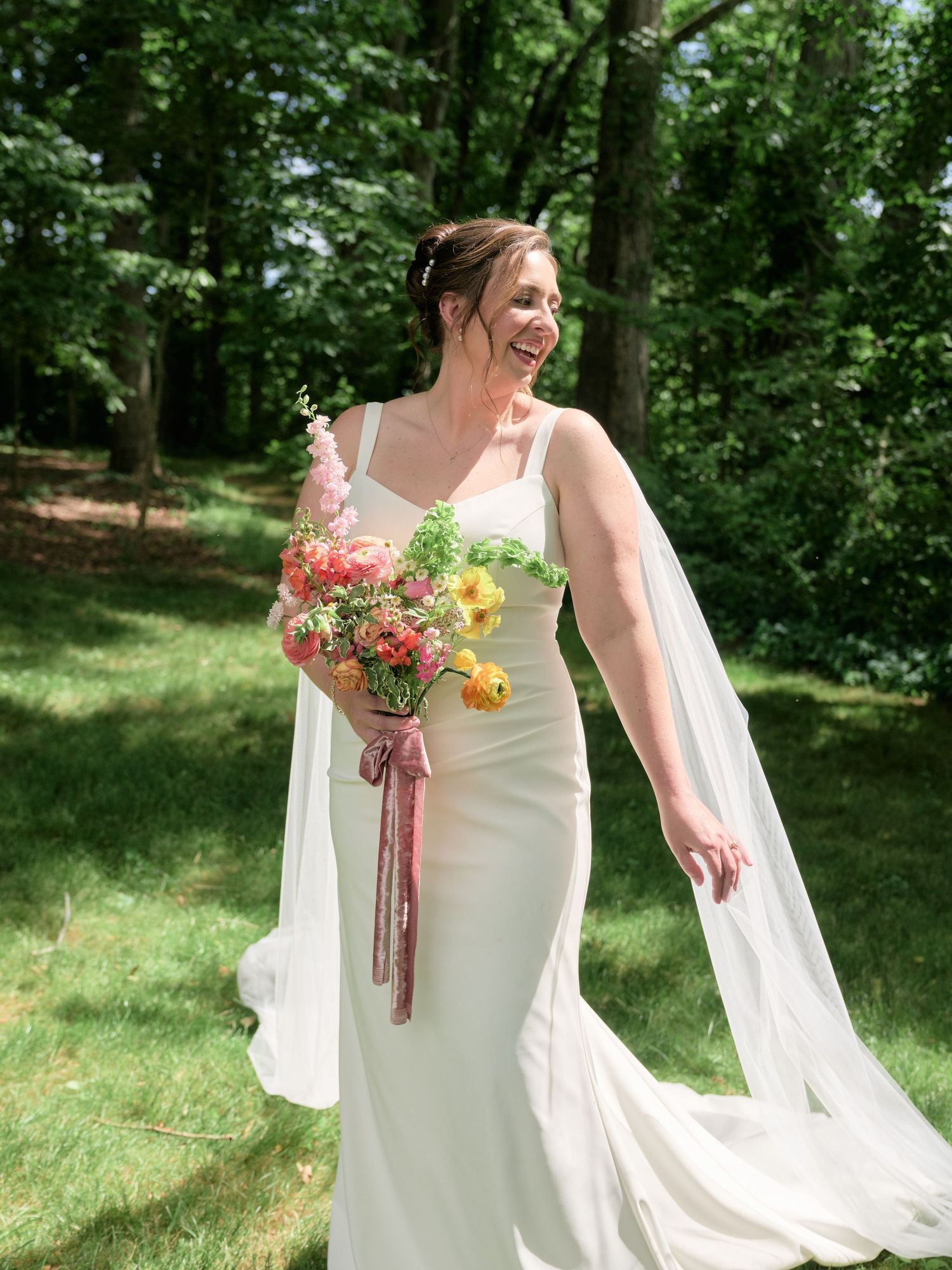 A bride in a fitted white gown walks across grass holding a colorful bouquet with streaming ribbons.