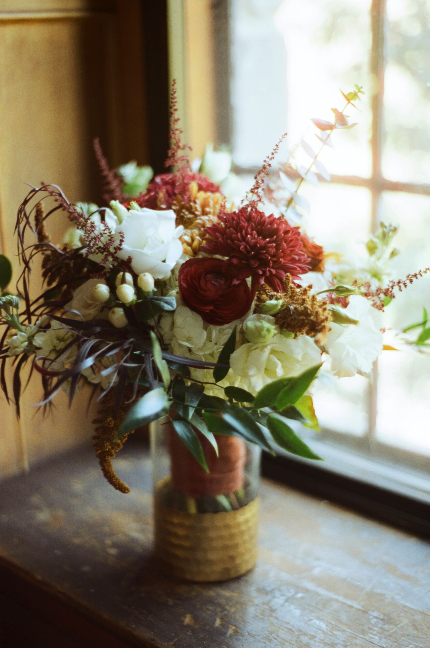 Romantic floral arrangement with white and burgundy flowers in a vintage vase by window.