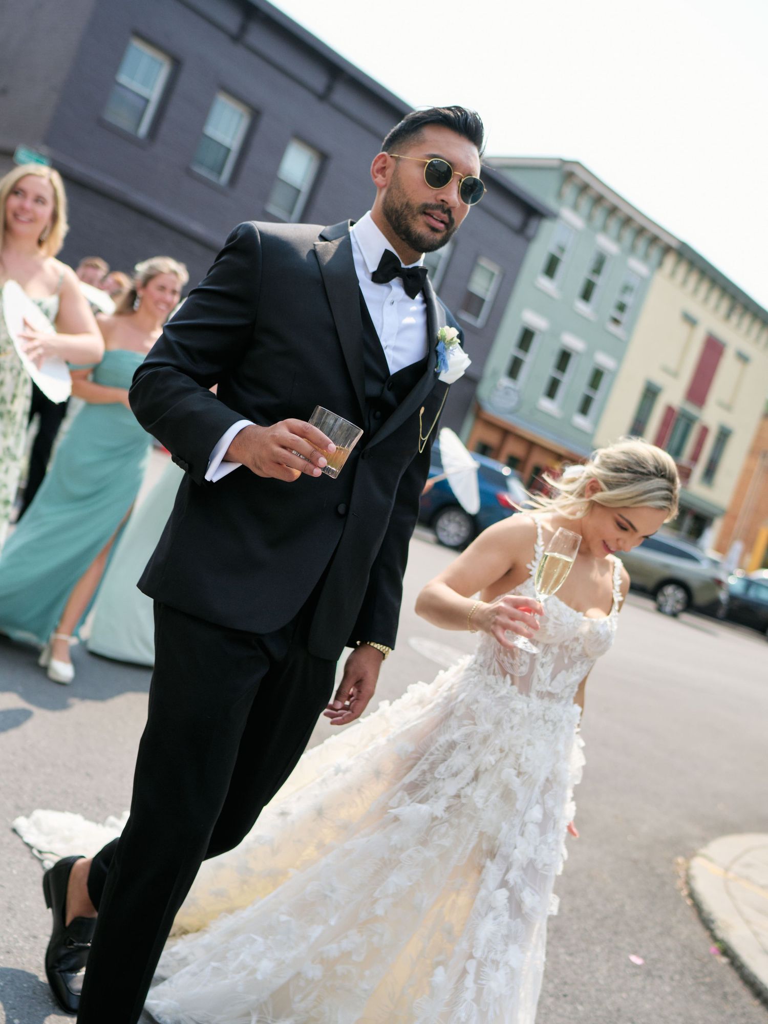 A joyful wedding couple laughs together on a city street with bridesmaid in green dress visible in background.