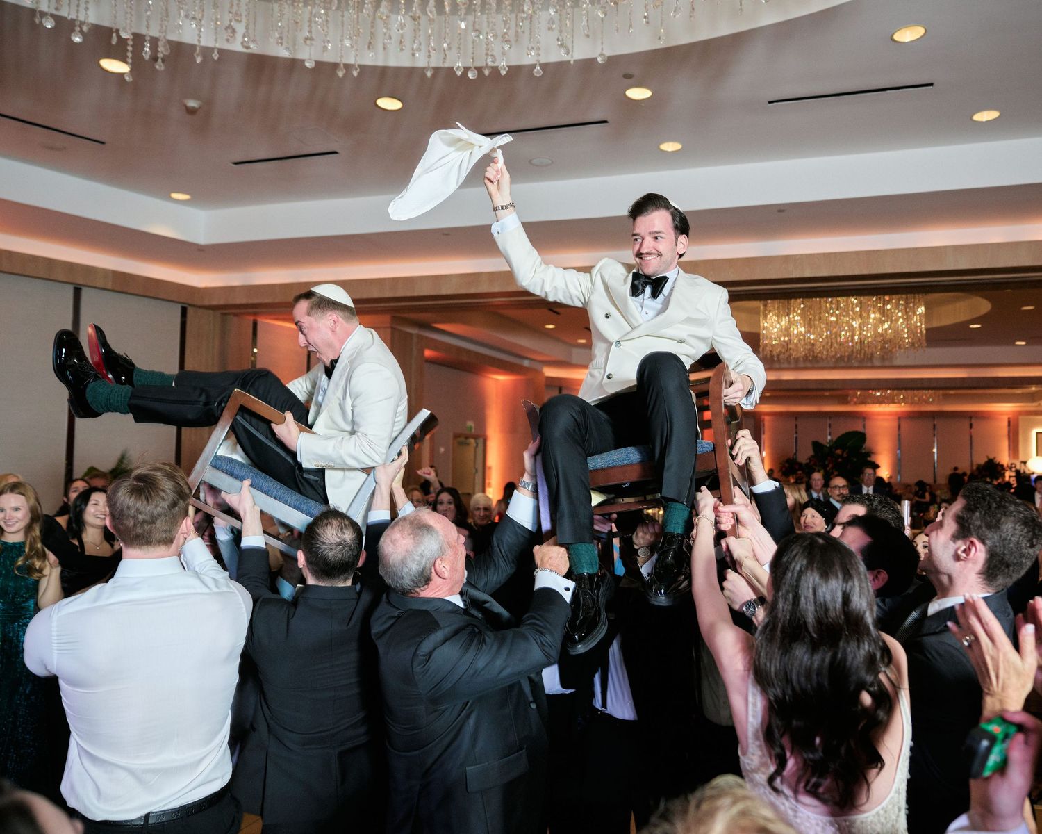 Wedding guests celebrate during the reception by lifting two people on chairs in a traditional dance celebration at a banquet hall.