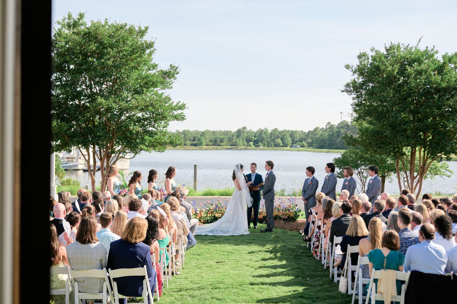 Outdoor wedding ceremony by a lakeside with guests seated on white chairs and trees framing the scenic view.