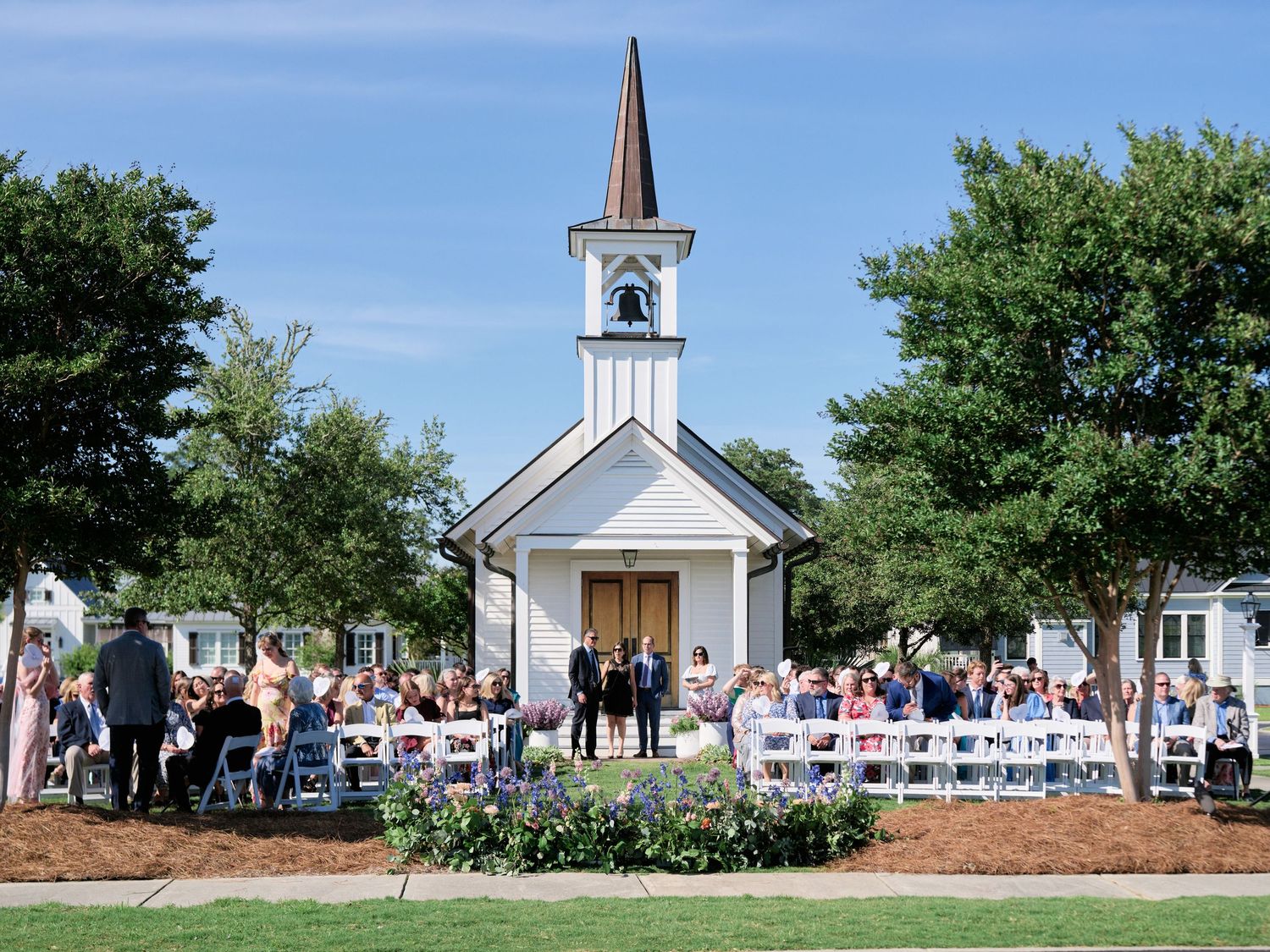A quaint white chapel with a steeple stands surrounded by guests at an outdoor wedding ceremony on a sunny day.